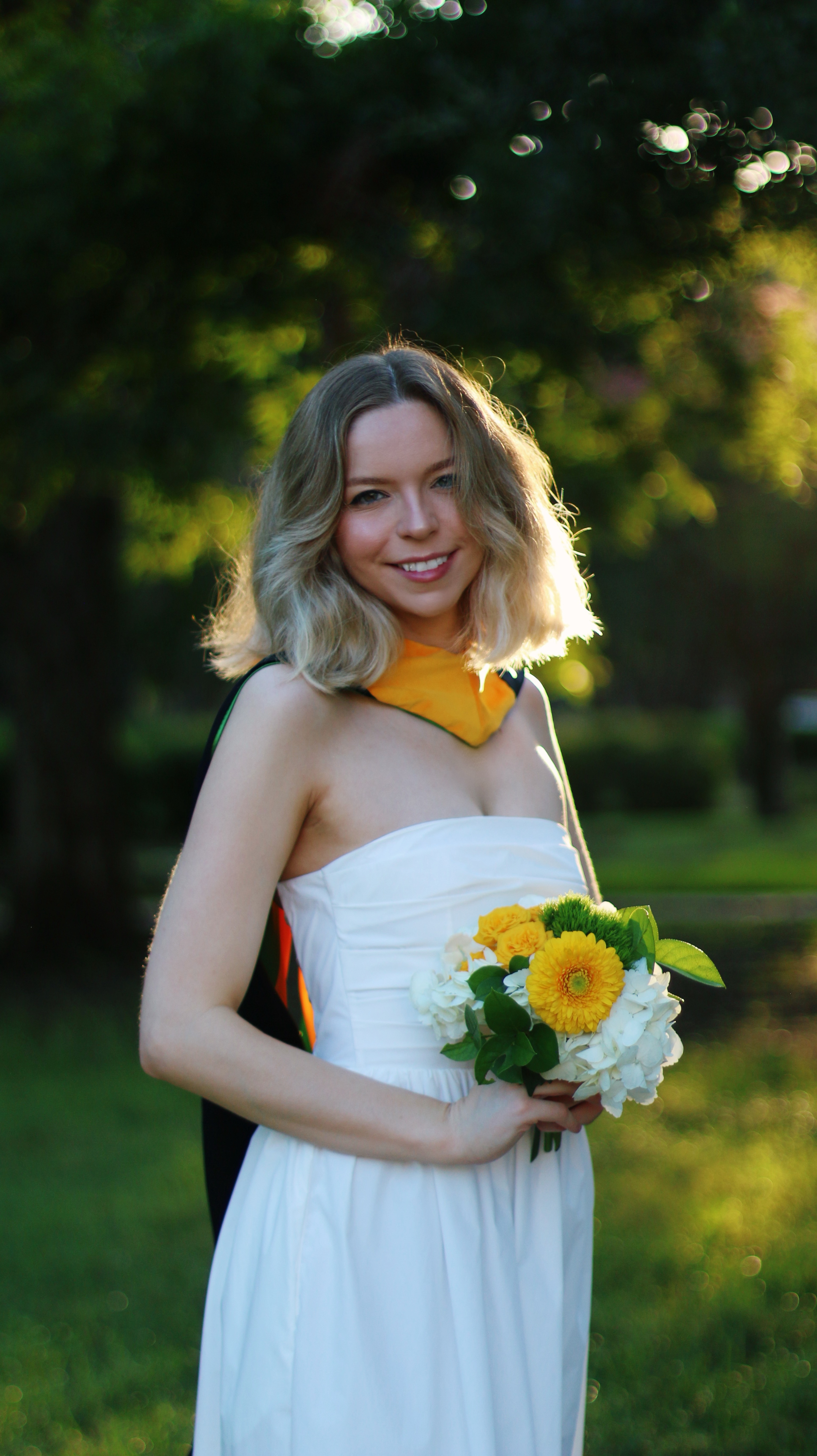 Baylor Graduation in white dress holding a bouquet of flowers in front trees