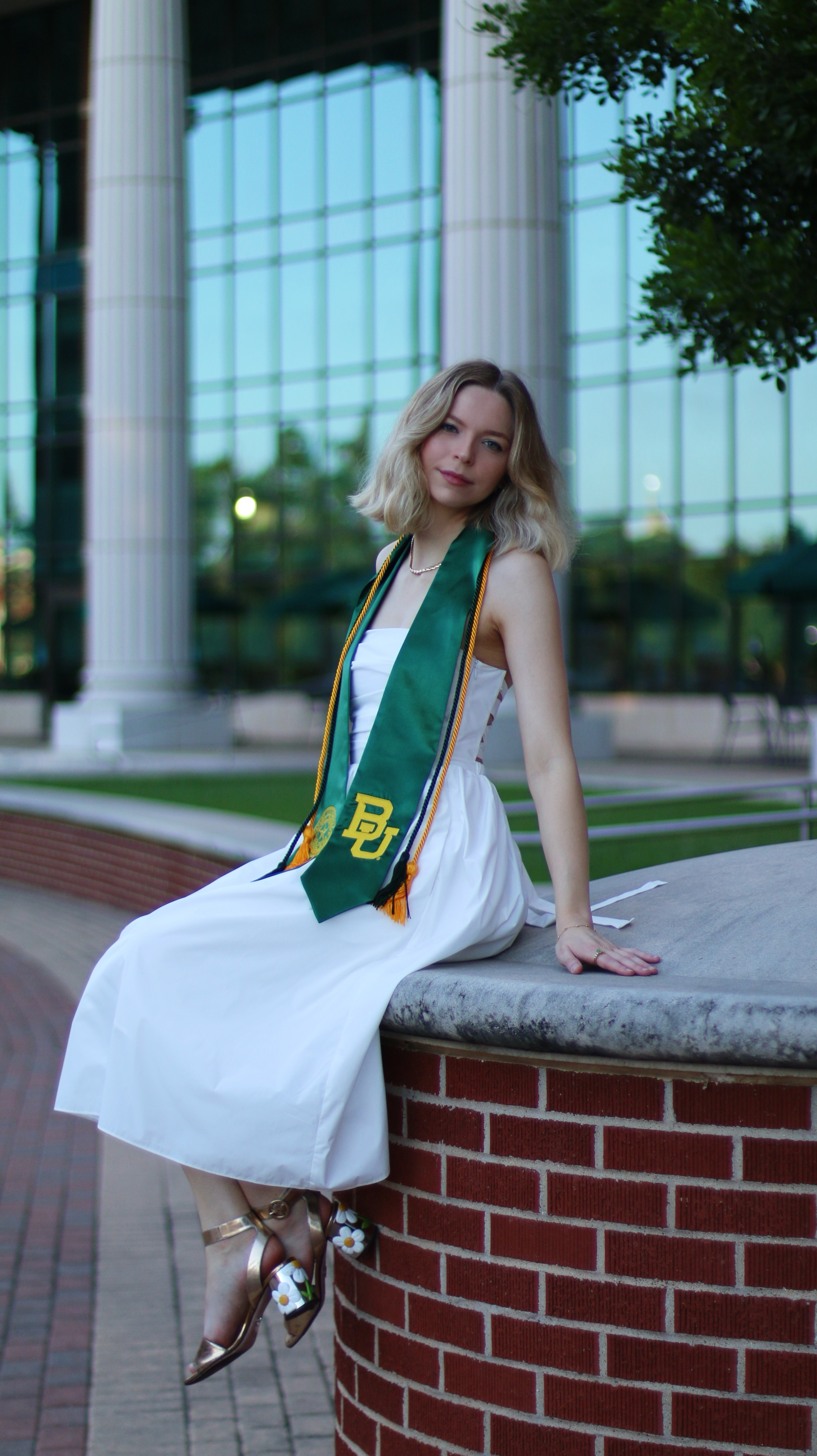 Baylor Graduation in white dress wearing sash and cords while sitting on a fountain