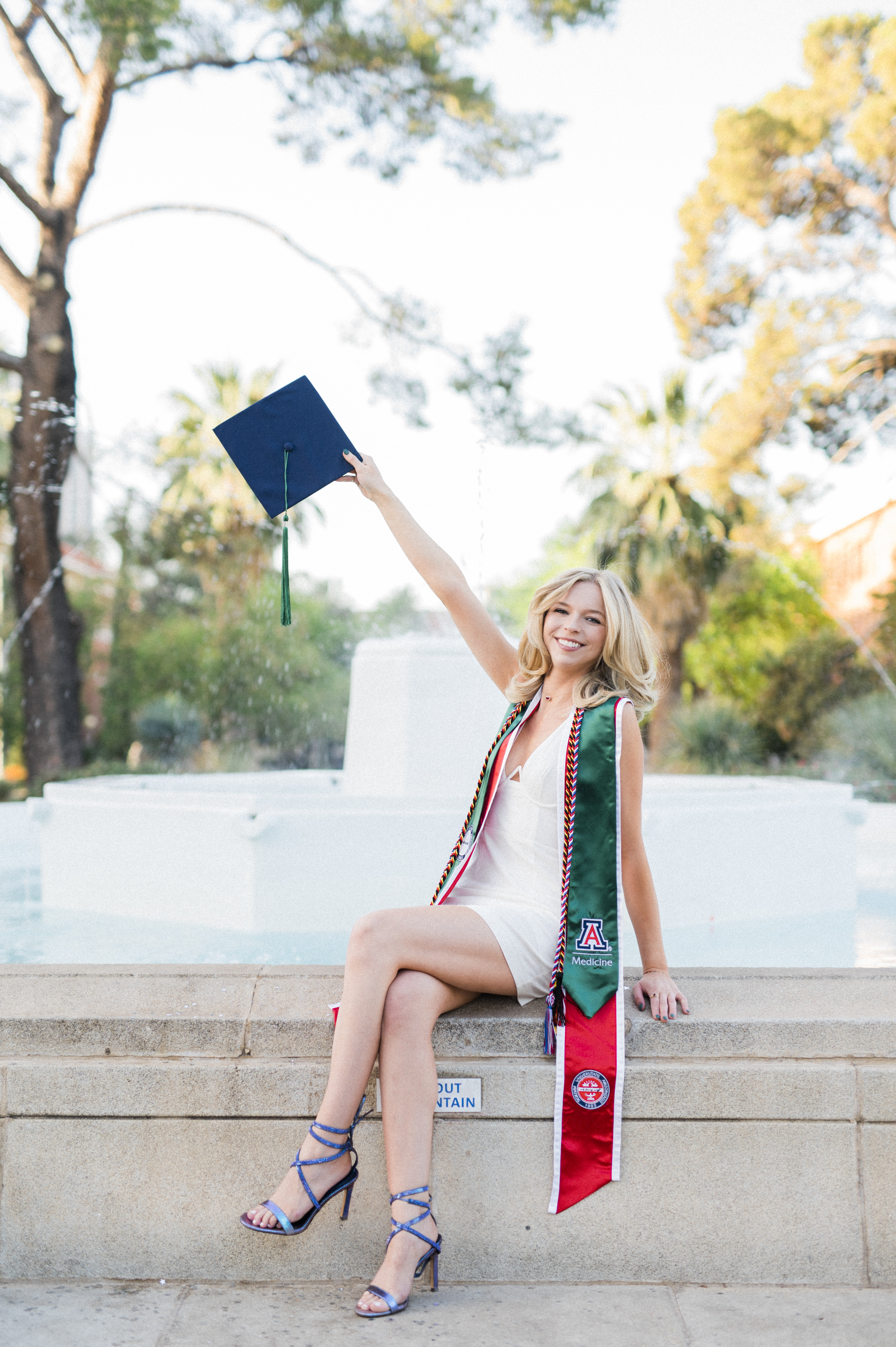 University of Arizona Graduation Picture with white dress sitting on a fountain with the unveristy sash on and cap lifted in the air