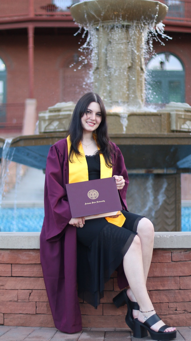 Graduate in maroon cap and gown holding Arizona State University diploma in front of a fountain