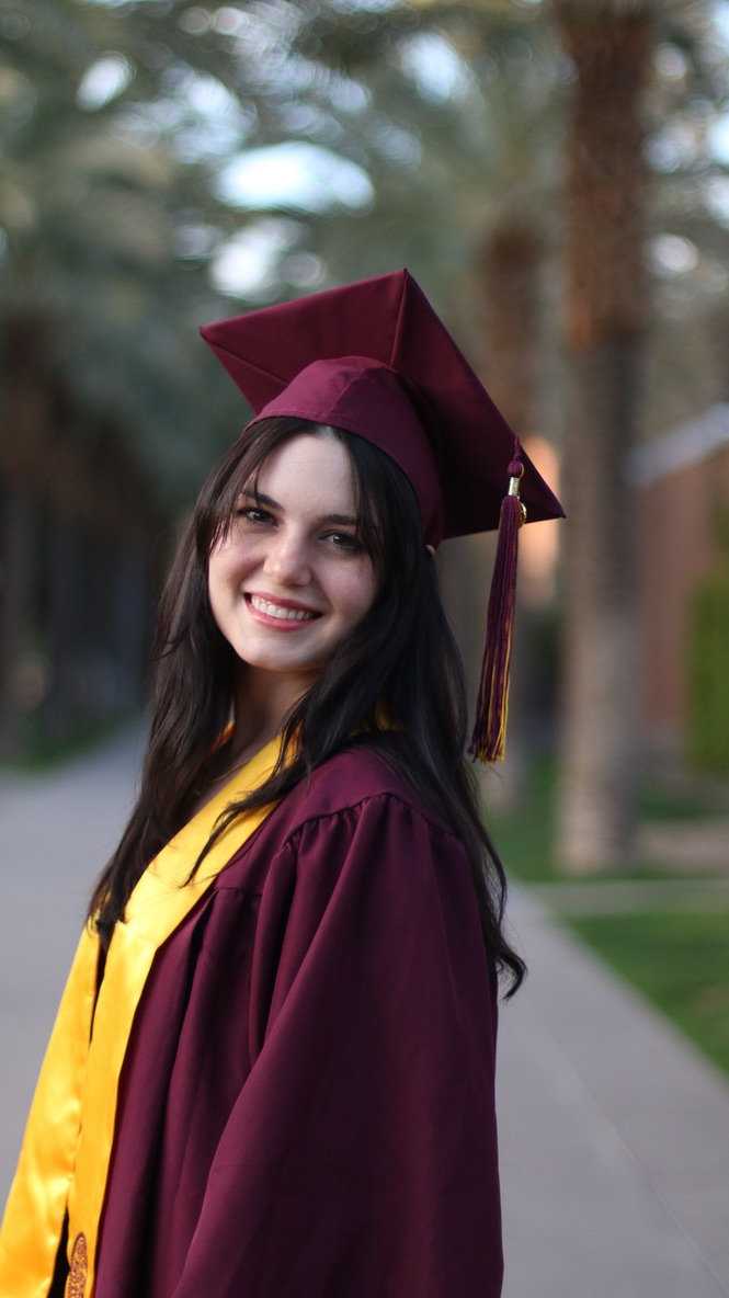 Smiling graduate in maroon cap and gown walking along a palm tree lined path
