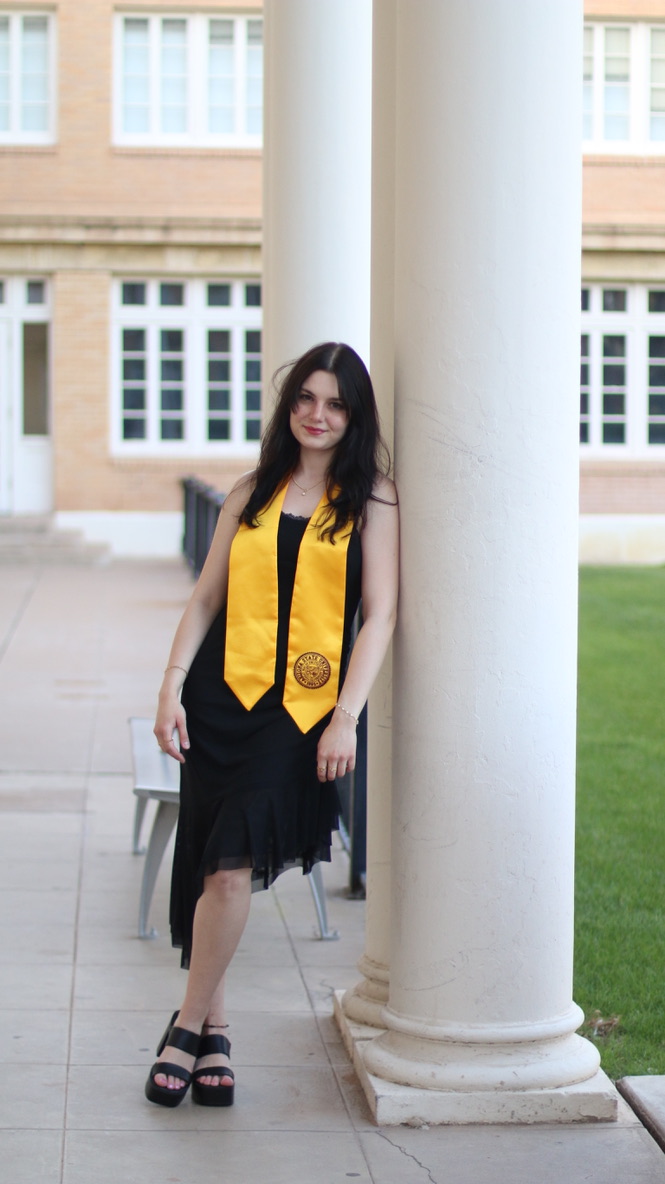 Graduate in gold sash leaning against a white column at a campus building