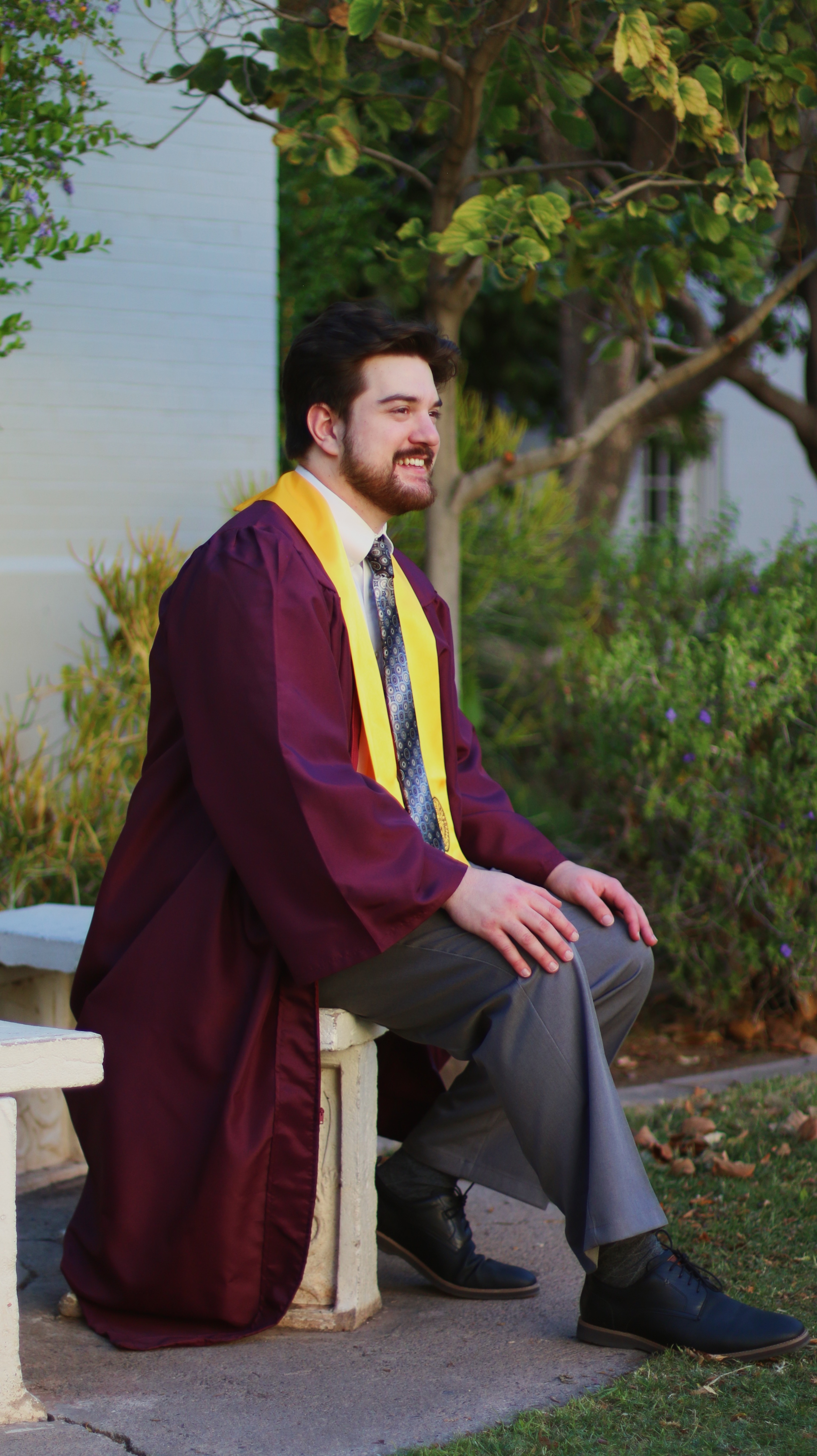 Male graduate in maroon gown and tie sitting on a bench in a garden setting