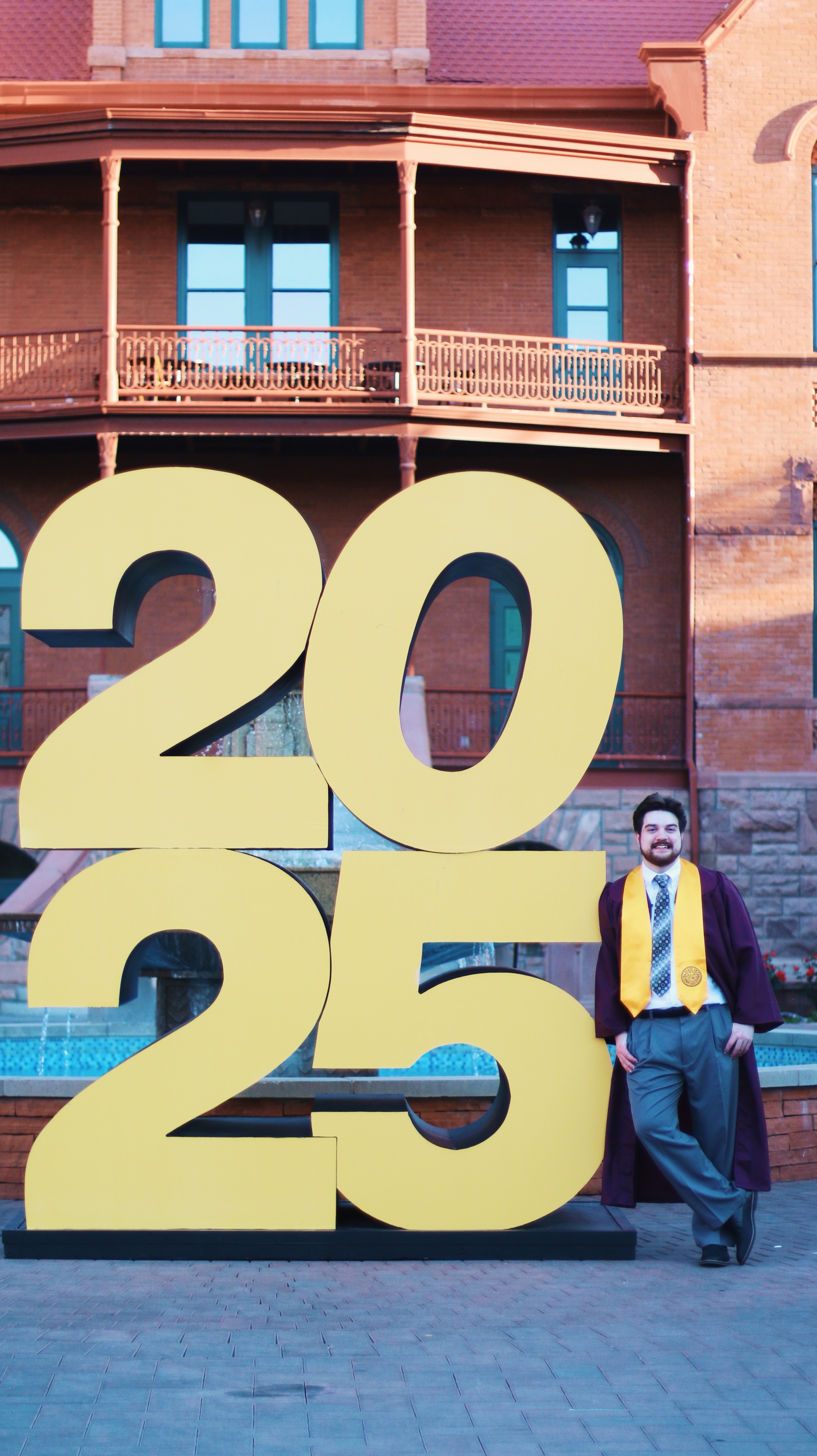 Male graduate leaning against a large 2025 sign in front of a historic brick building