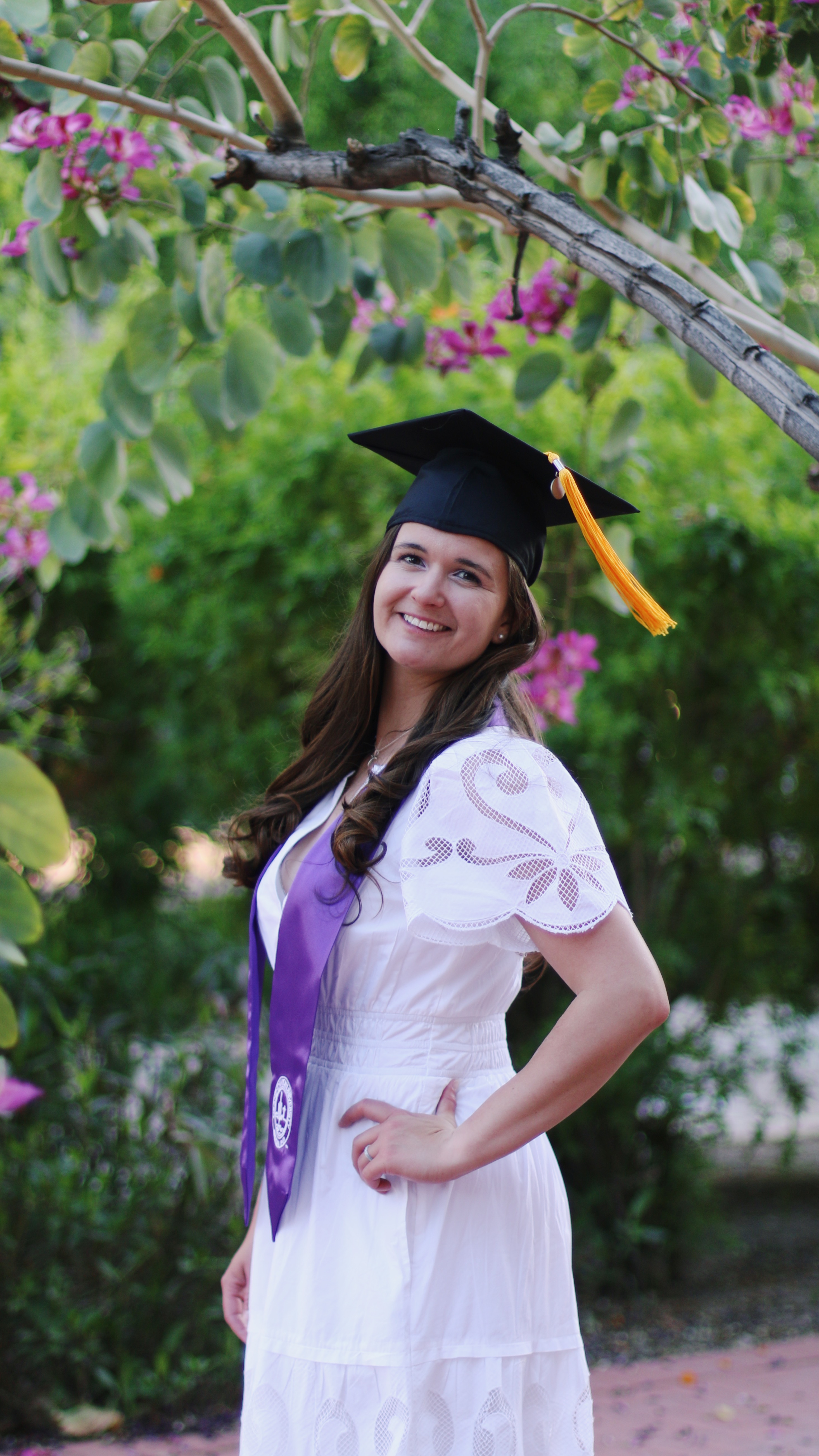 Smiling graduate in black cap and purple sash standing among pink flowering trees