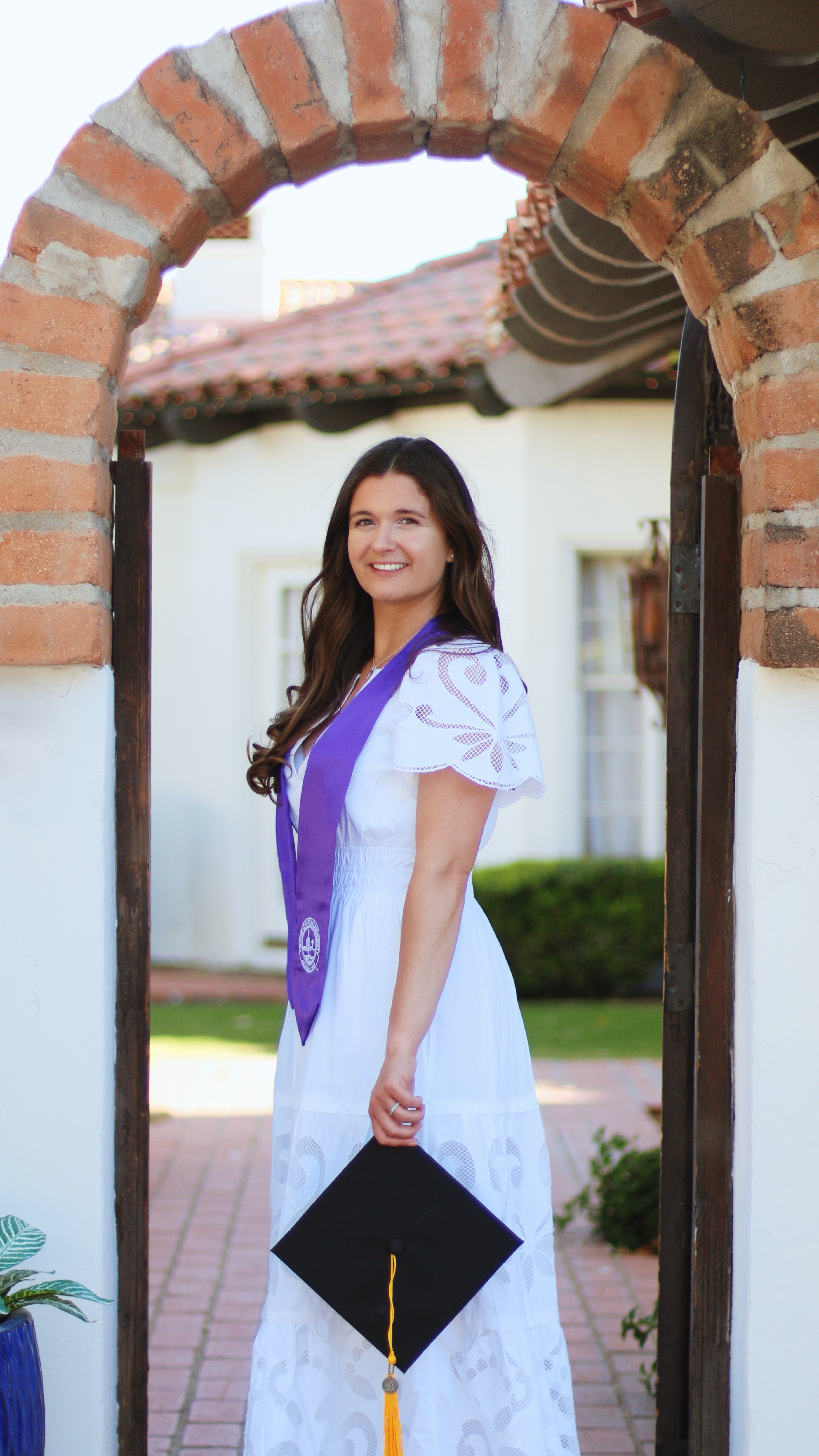 Graduate in white dress and purple sash standing in a brick archway doorway