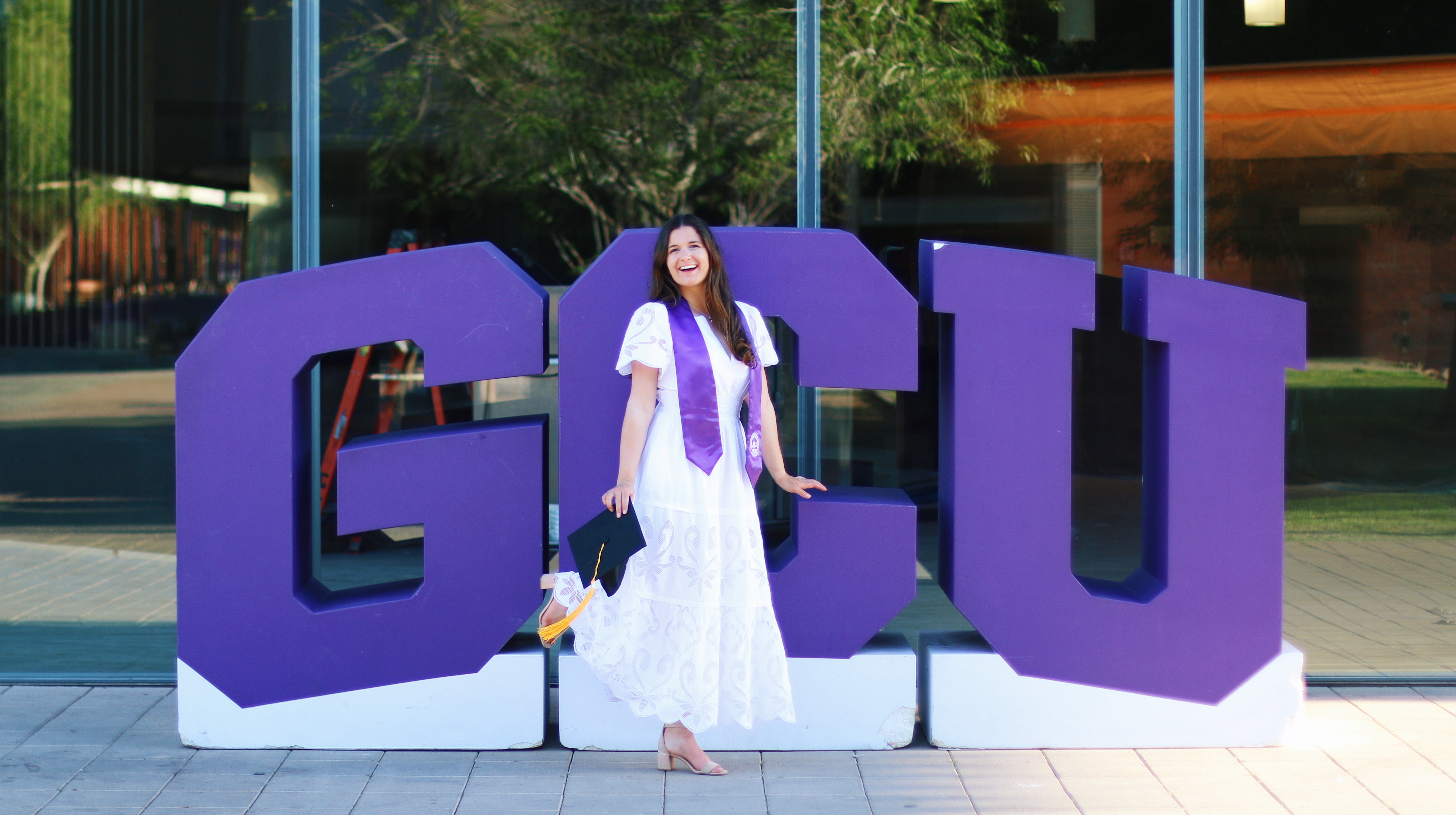 Graduate in white dress and purple sash laughing in front of large GCU block letters