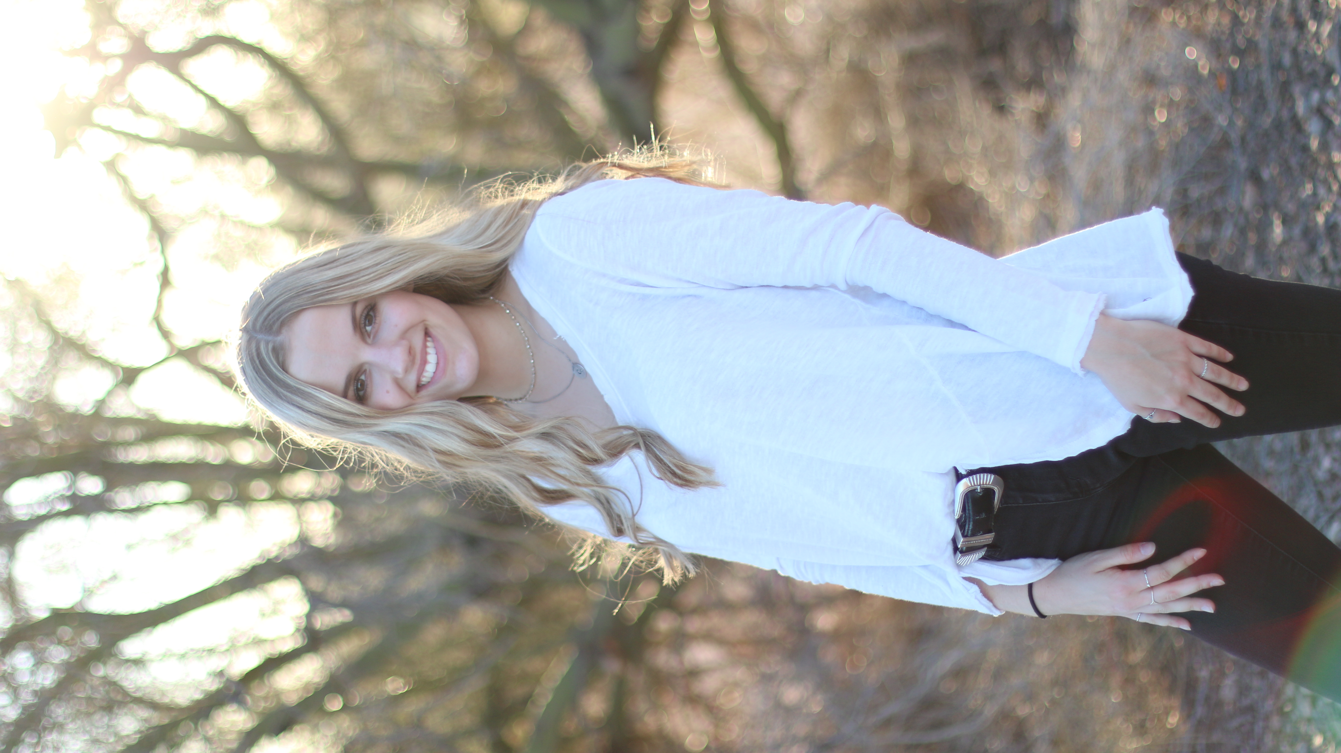 Young woman with blonde hair smiling in a sunlit wooded trail wearing a white top