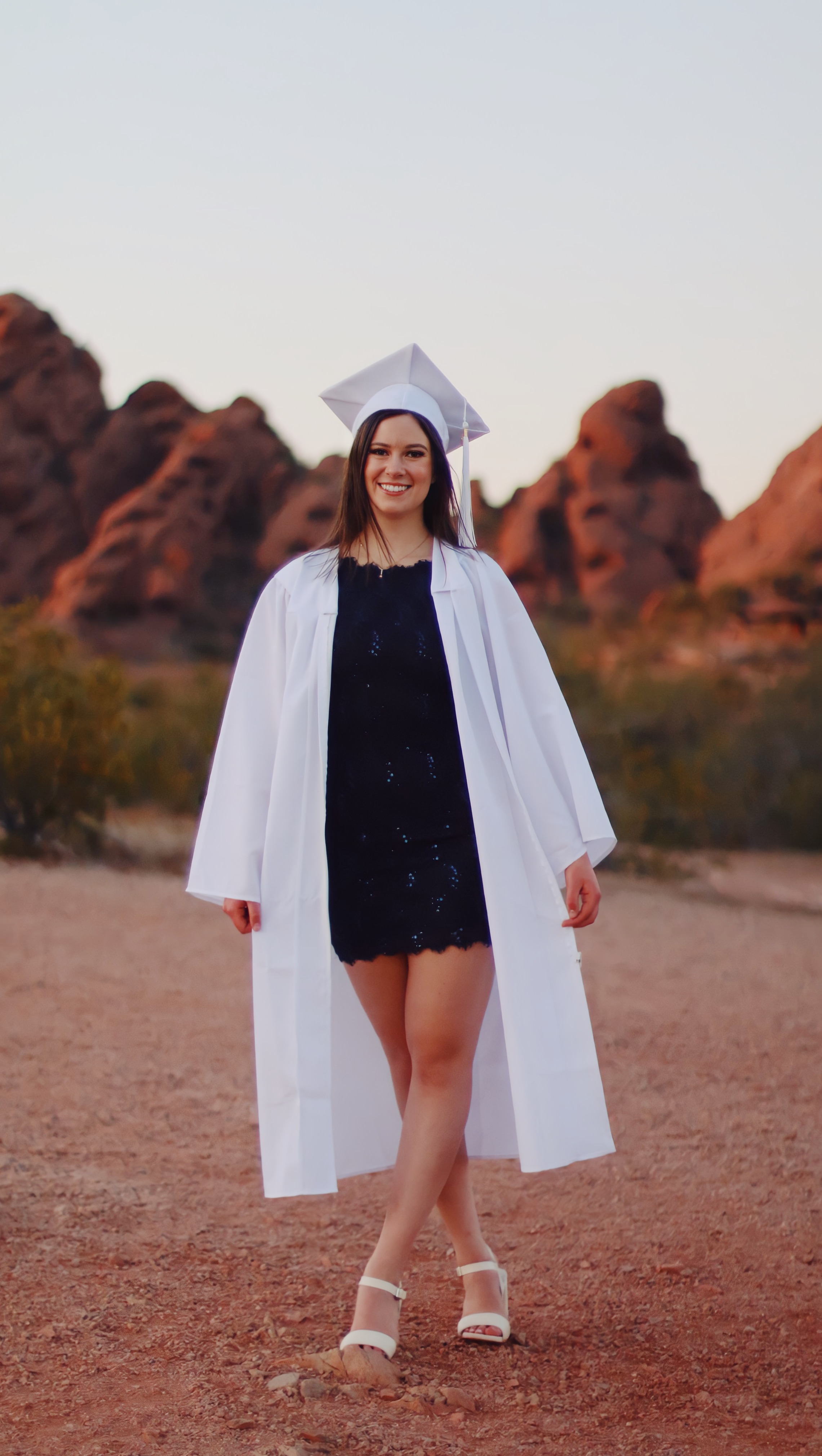 Graduate in white cap and gown walking at Papago Park with red rock formations in the background