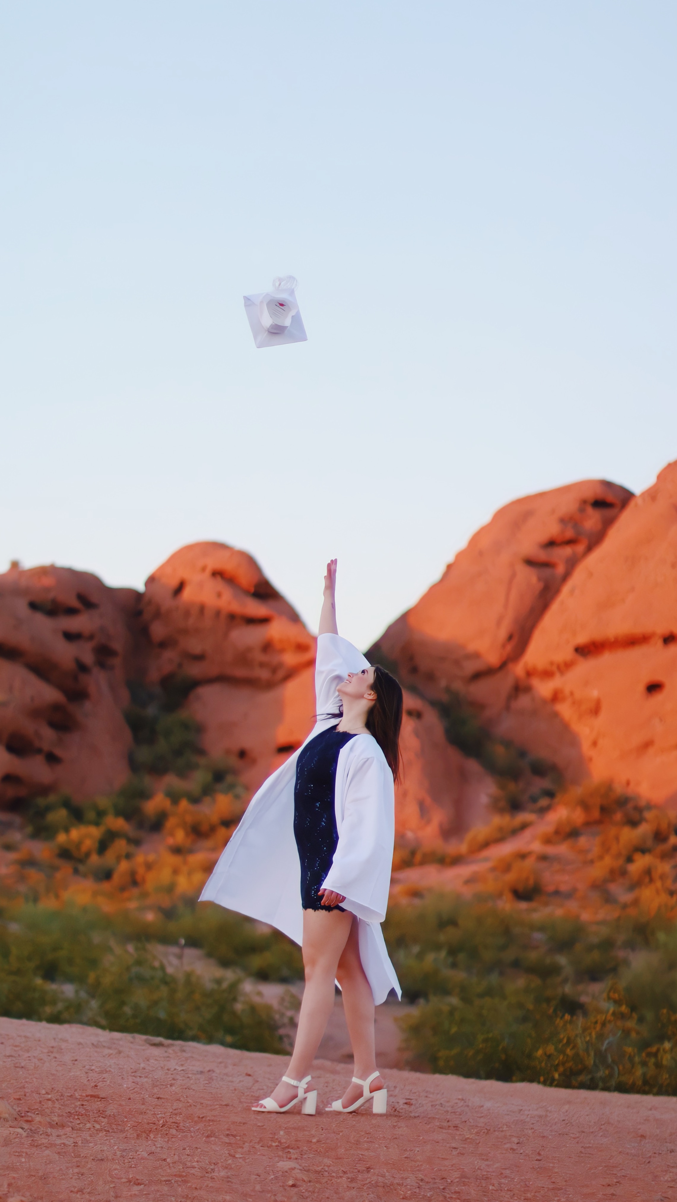 Graduate in white gown tossing her cap into the air at Papago Park at golden hou