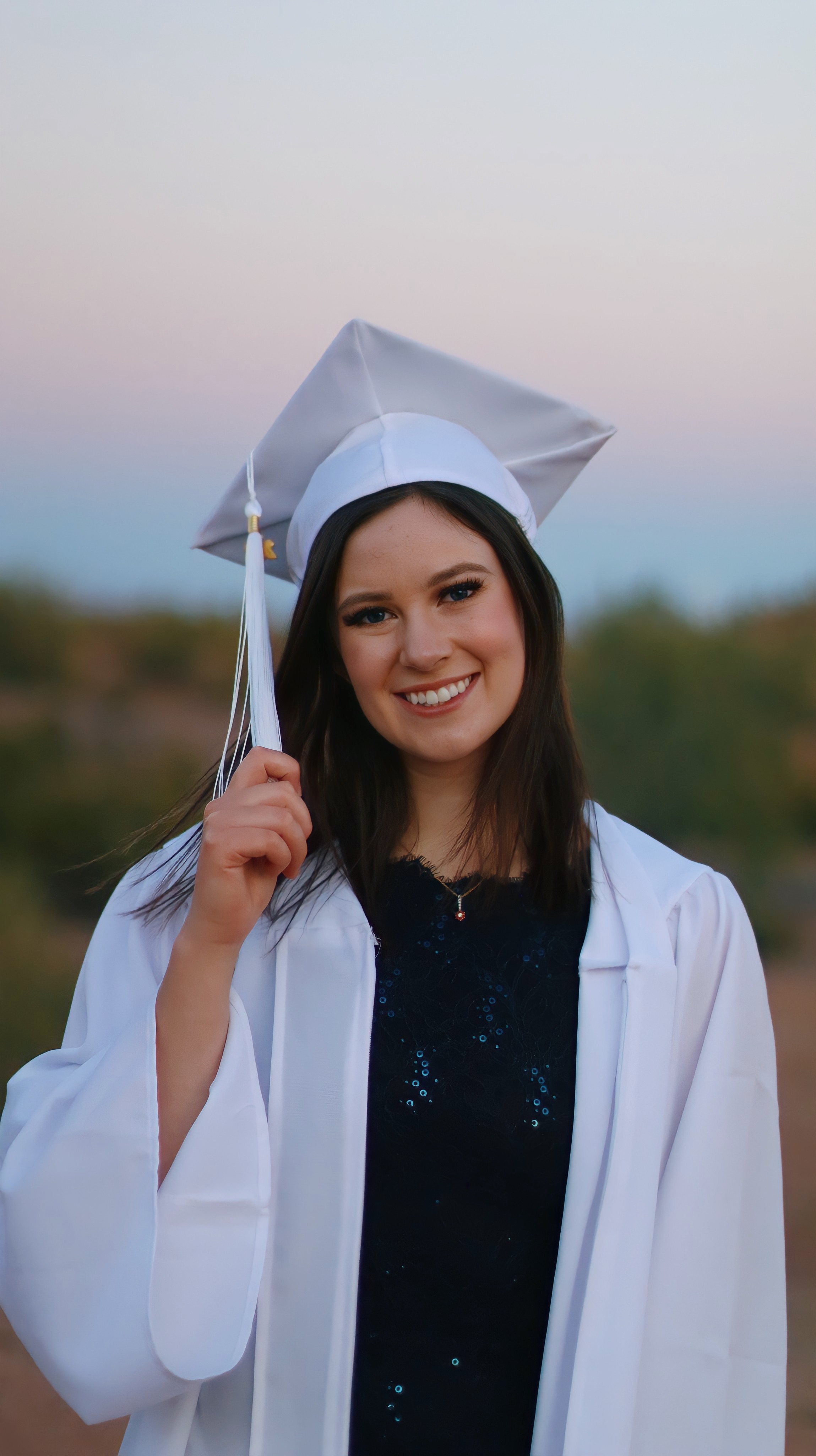 Graduate in white cap and gown holding tassel and smiling at Papago Park