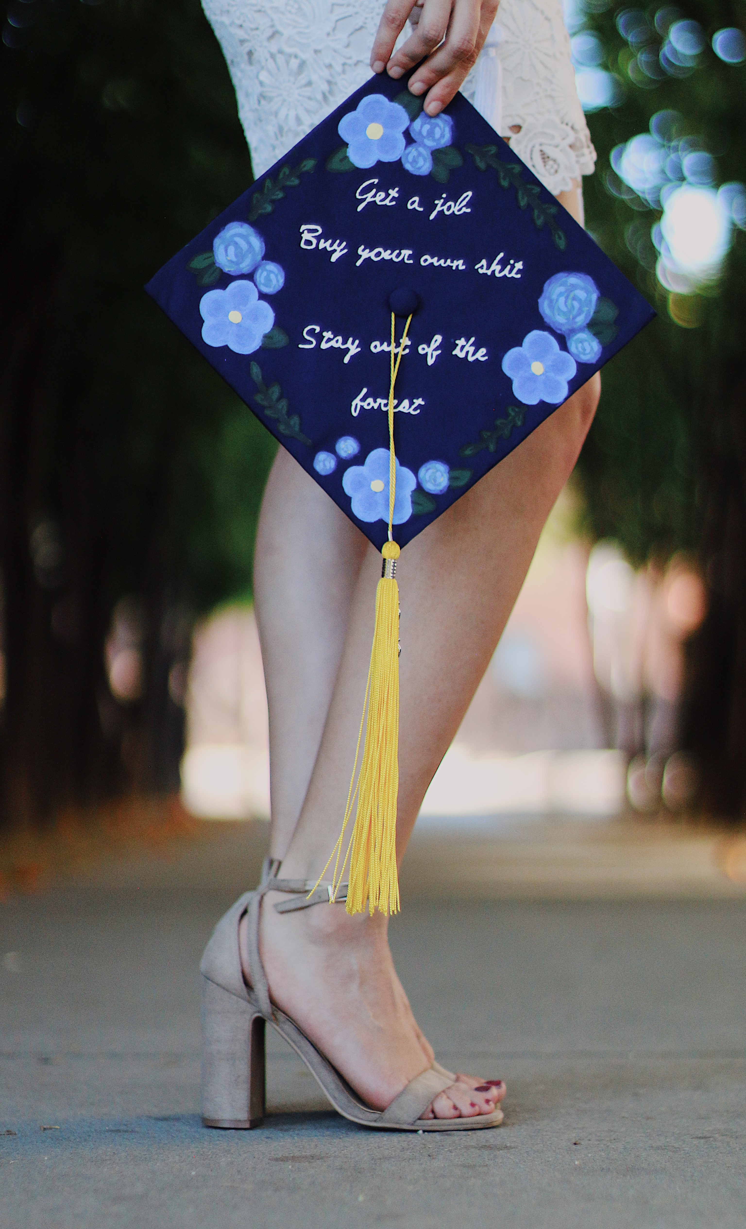 Close up of a decorated navy graduation cap with blue flowers and a humorous quote held at waist level