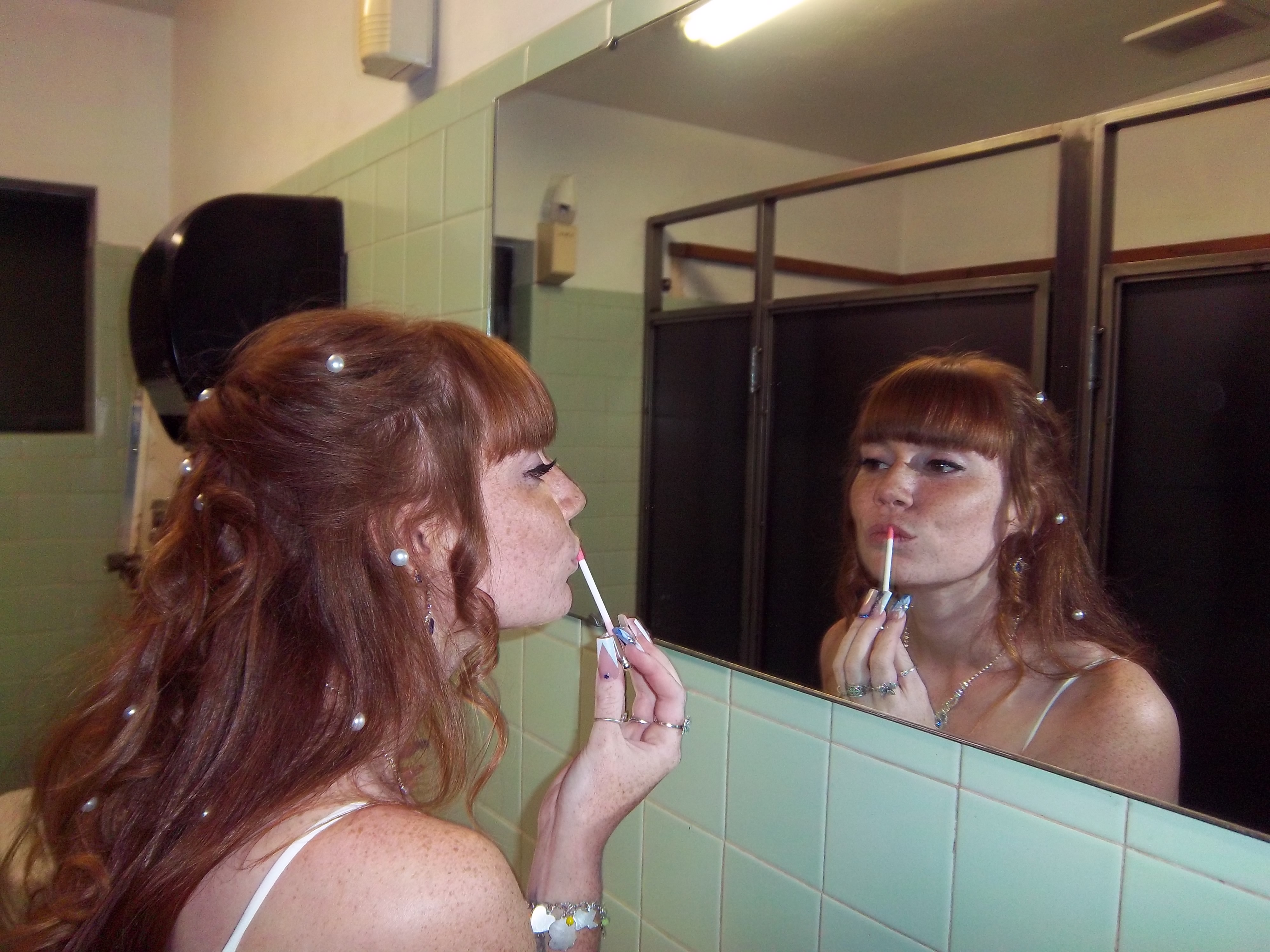 Young woman with red hair applying lip gloss in a bathroom mirror reflection