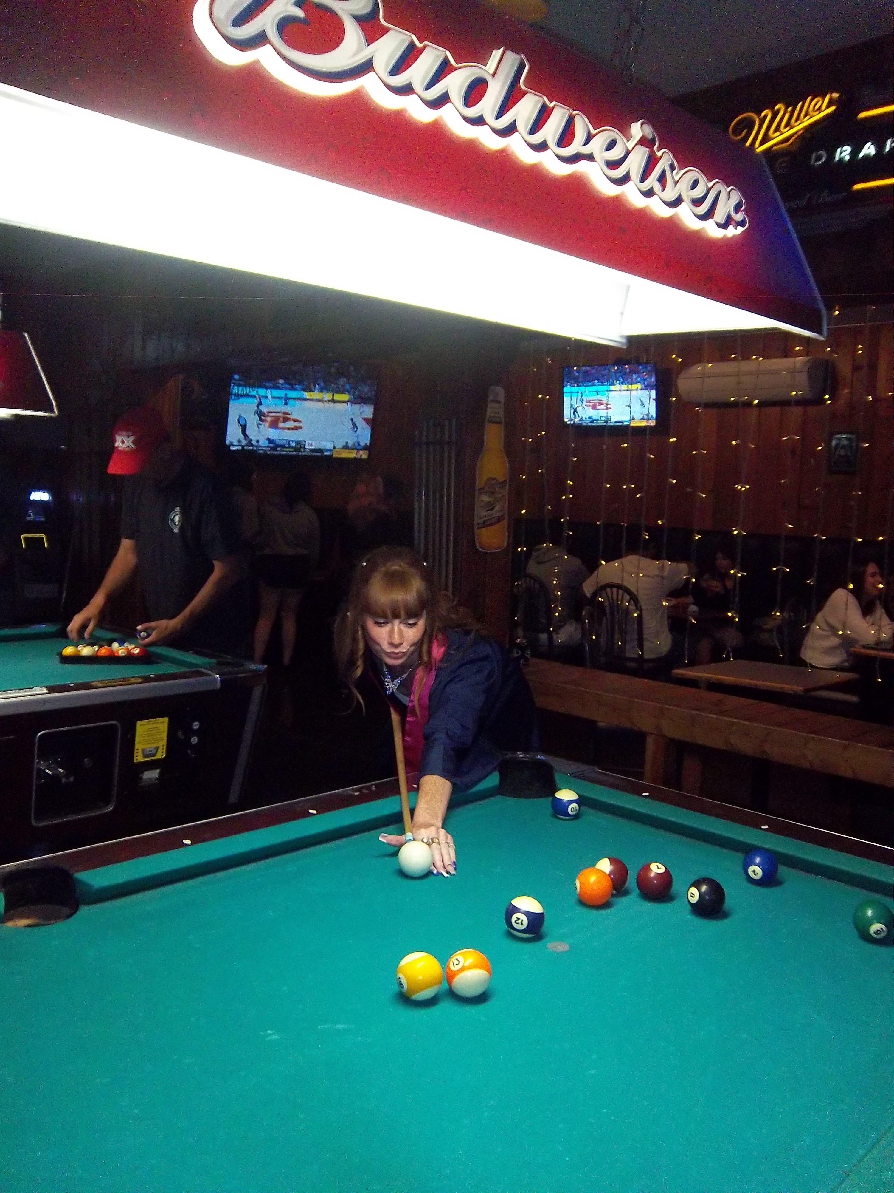 Woman with red hair leaning over a pool table taking a shot at a bar with neon signs