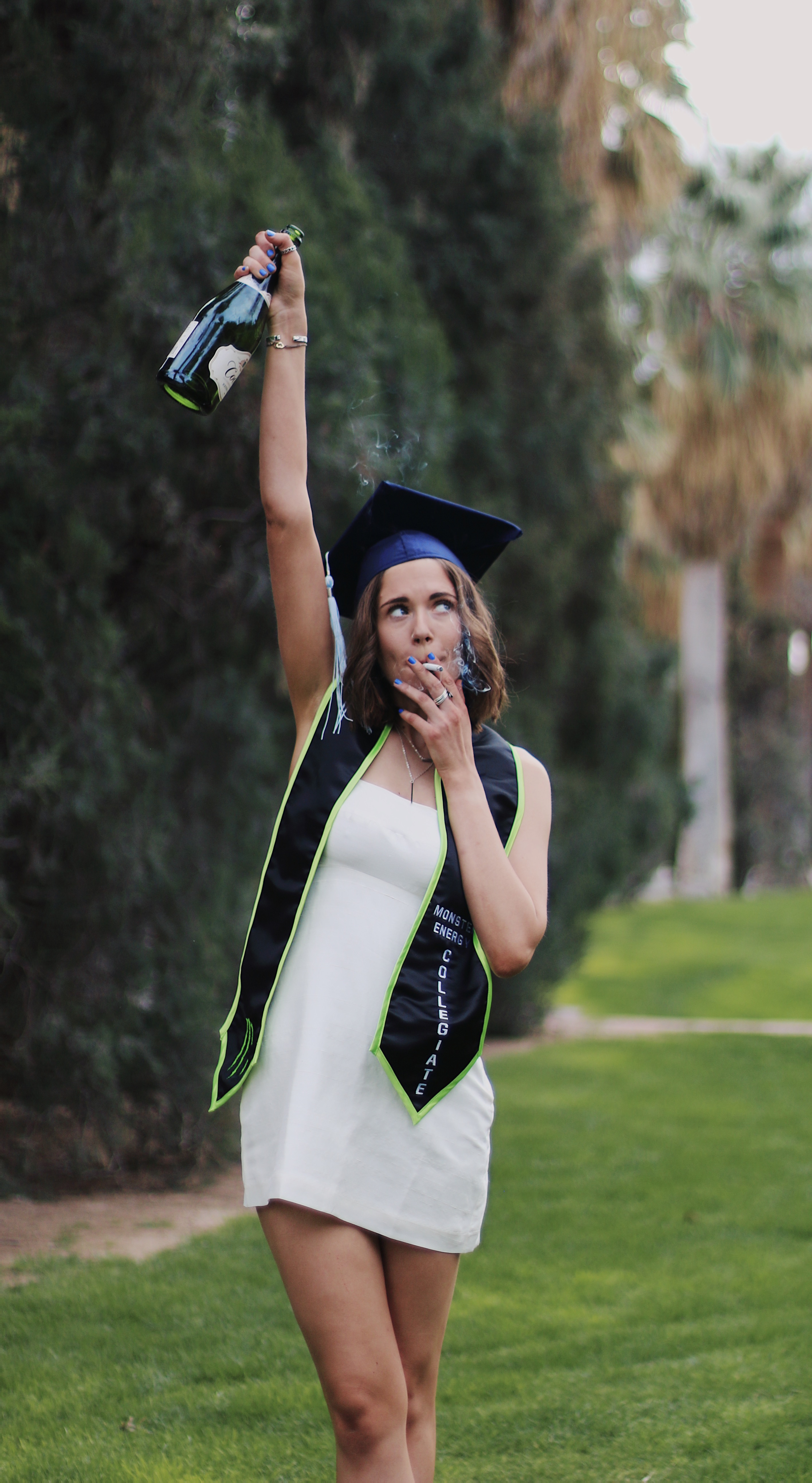 Graduate in navy cap and white dress holding champagne bottle above her head on a grassy campus lawn
