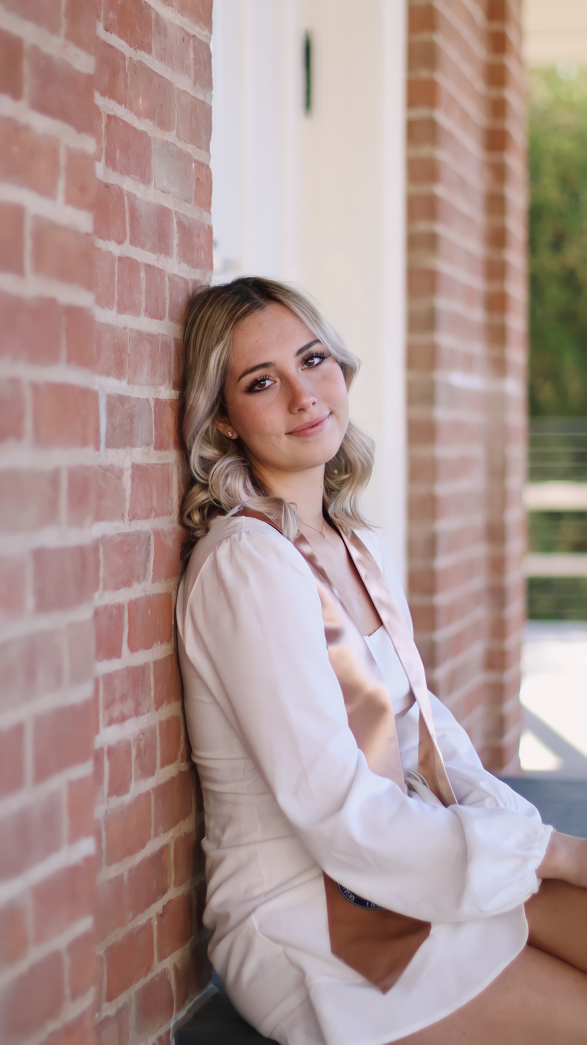 Young woman in white blazer leaning against a brick wall smiling softly at the camera