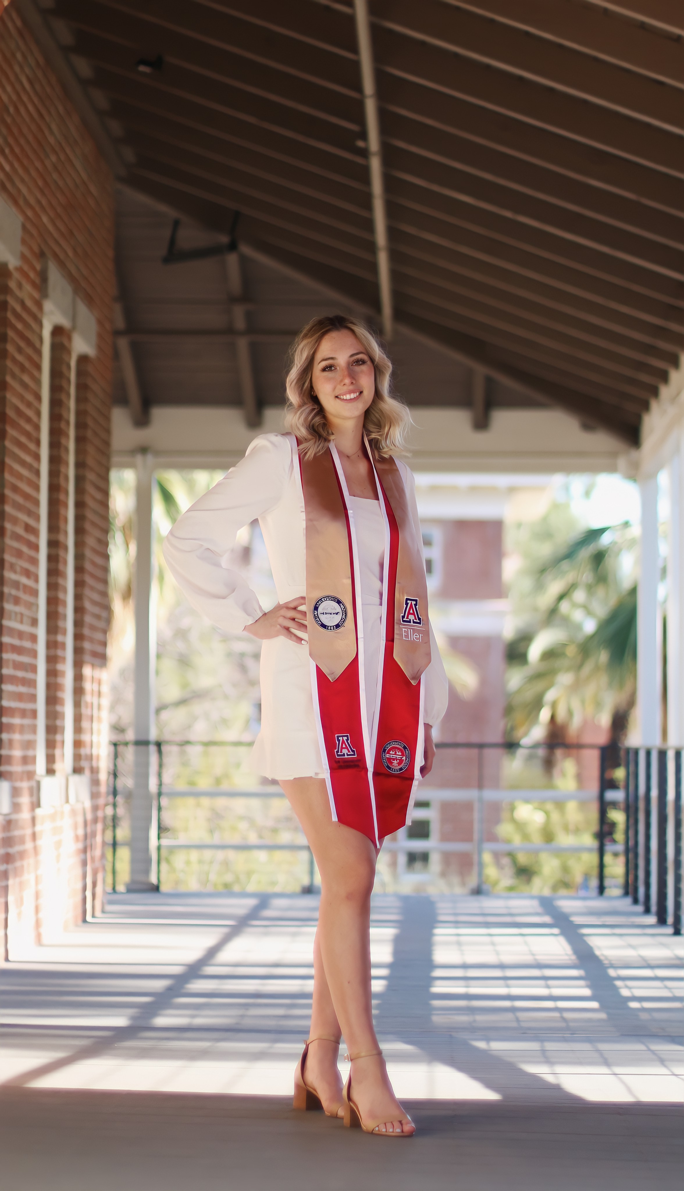 Graduate in white dress and University of Arizona red sash posing in a covered campus walkway