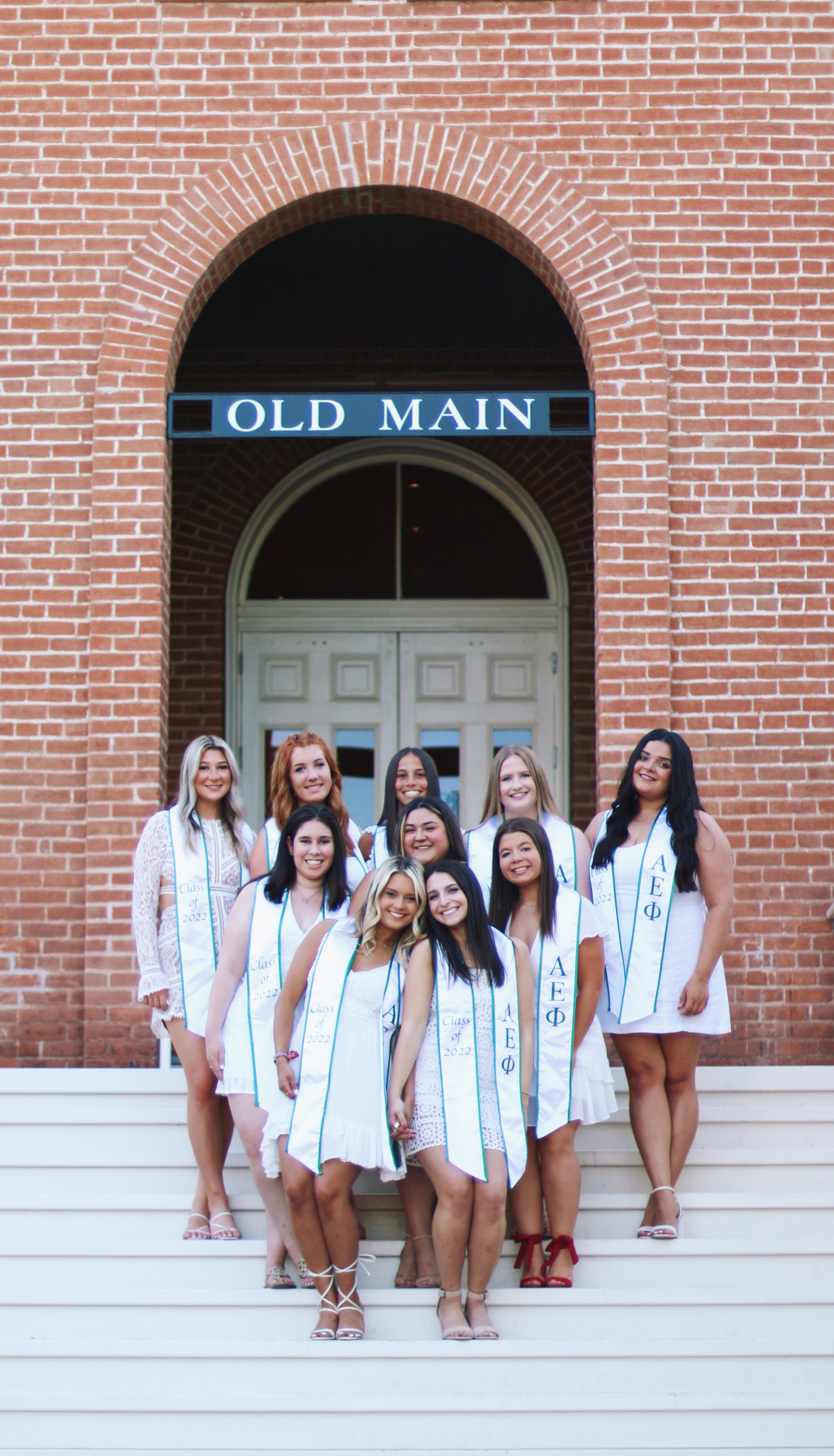 Group of nine women in white dresses with Alpha Epsilon Phi graduation sashes posing on the steps of Old Main
