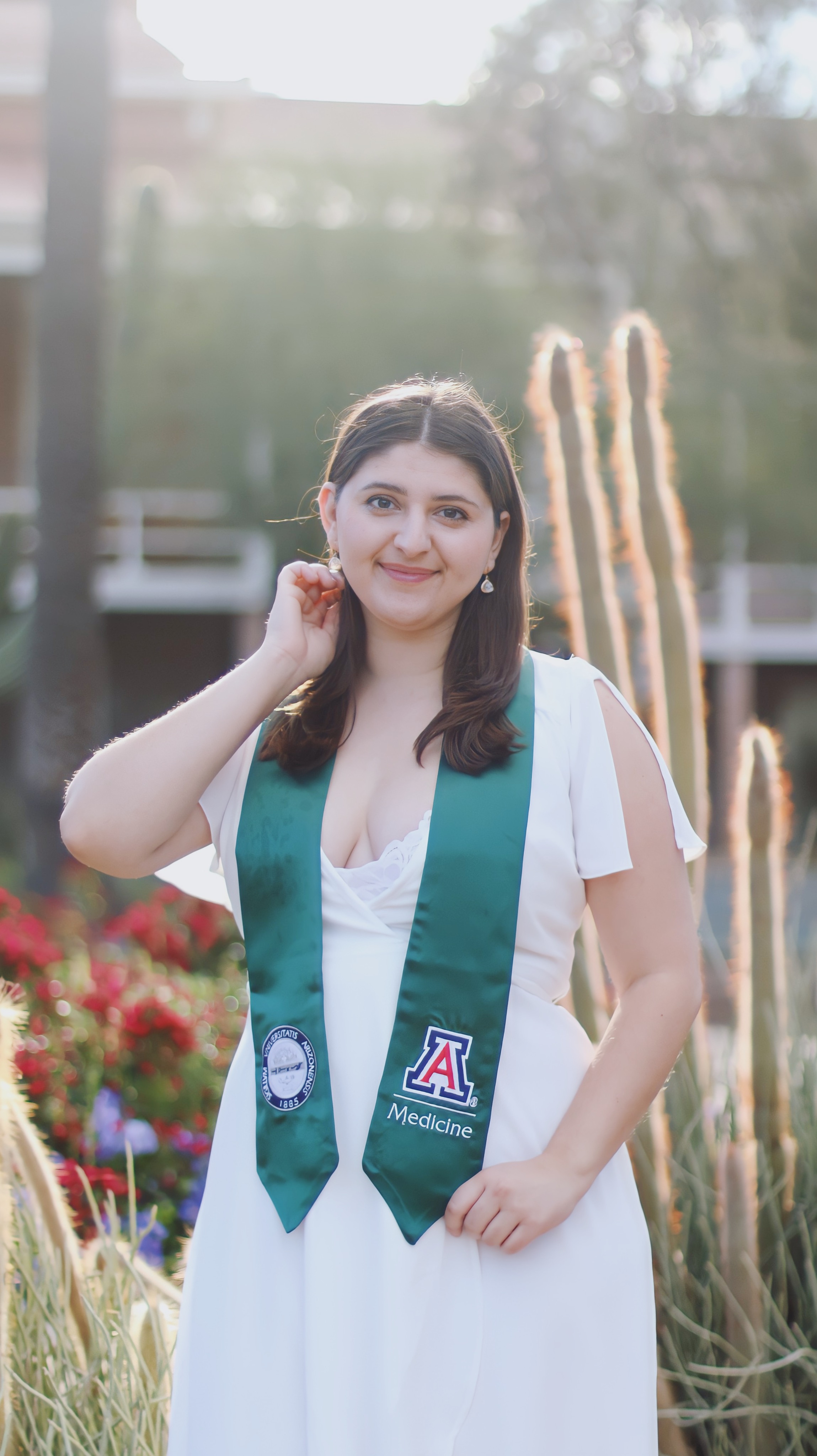 Graduate in white dress and teal University of Arizona Medicine sash standing among desert cacti and flowers at golden hour