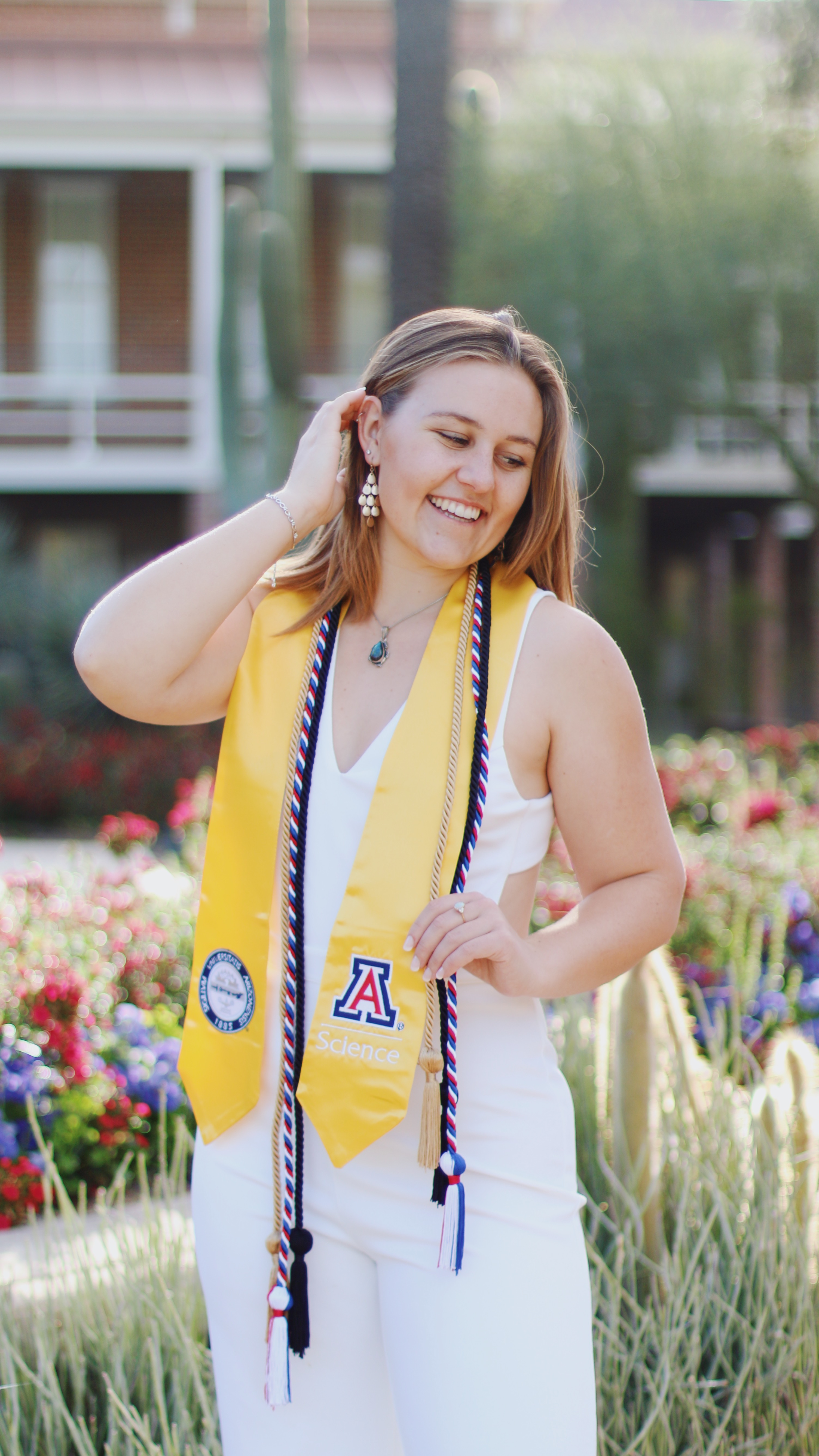 Smiling graduate in white outfit and yellow University of Arizona Science sash among red and purple flowers on campus
