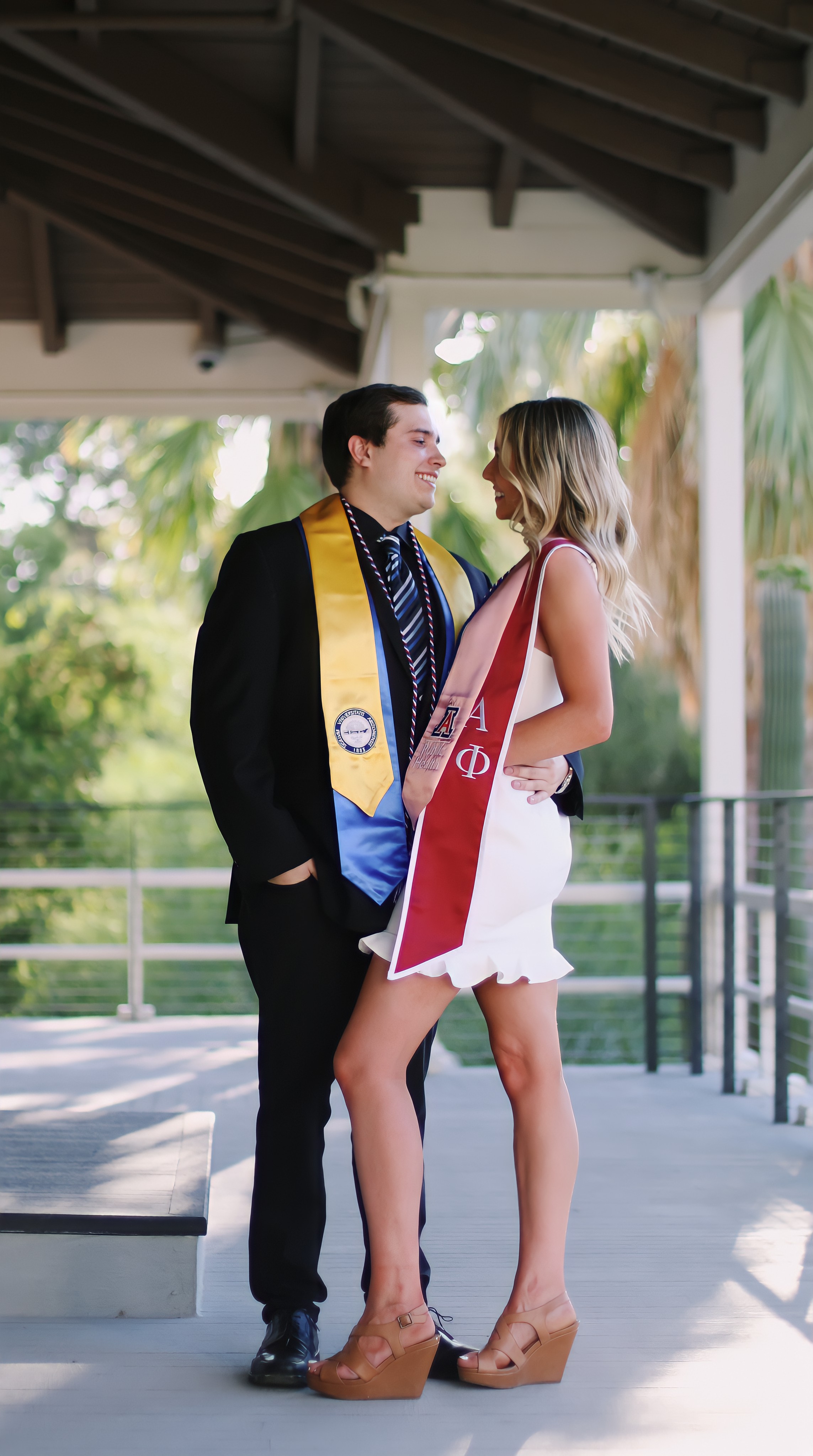 Male and female graduate in formal attire with University of Arizona sashes smiling at each other on a covered walkway