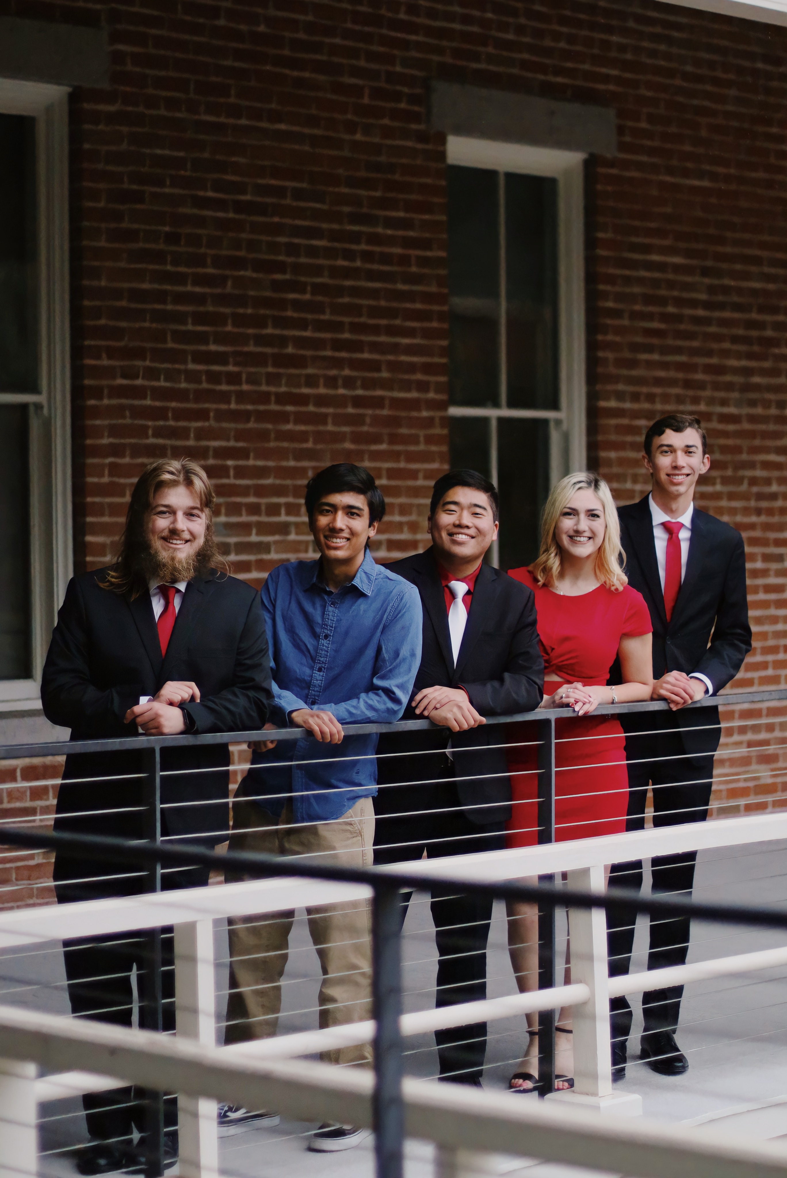 Group of five university students in formal attire leaning on a railing and smiling in front of a brick building
