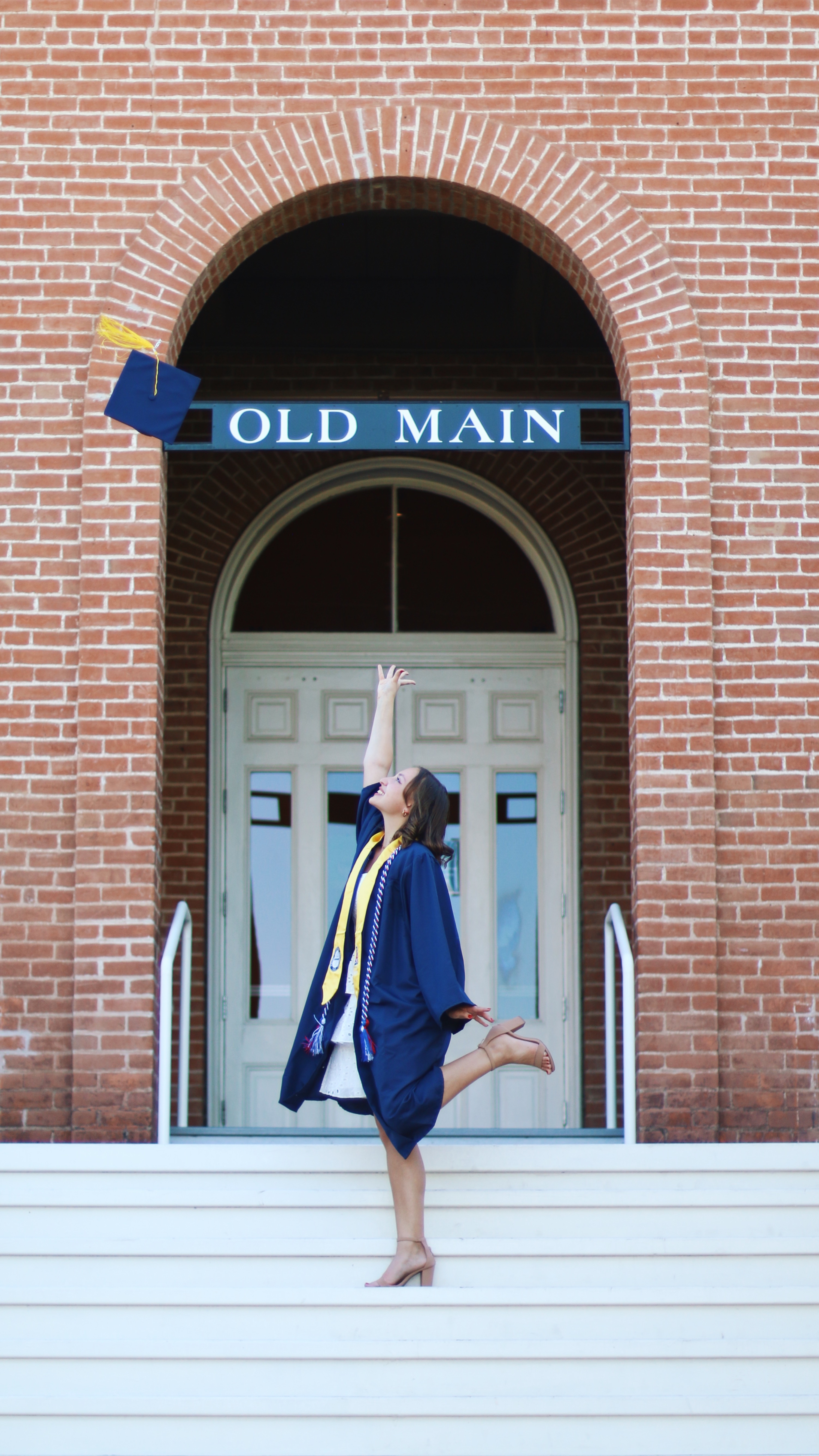 Graduate in navy cap and gown joyfully tossing her cap in front of Old Main building at the University of Arizona