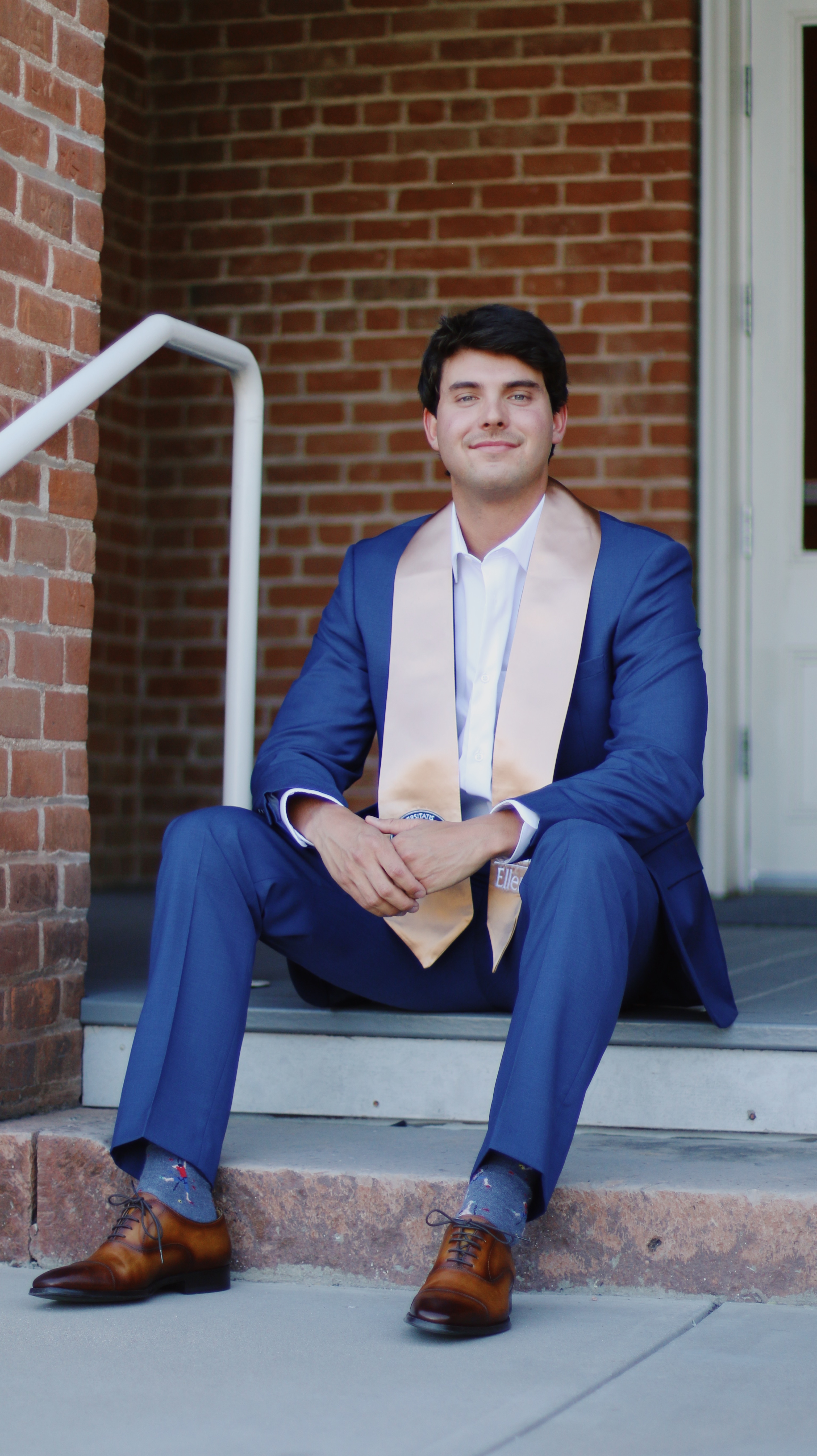 Young man in blue suit with graduation sash sitting on steps outside a brick building smiling at the camera