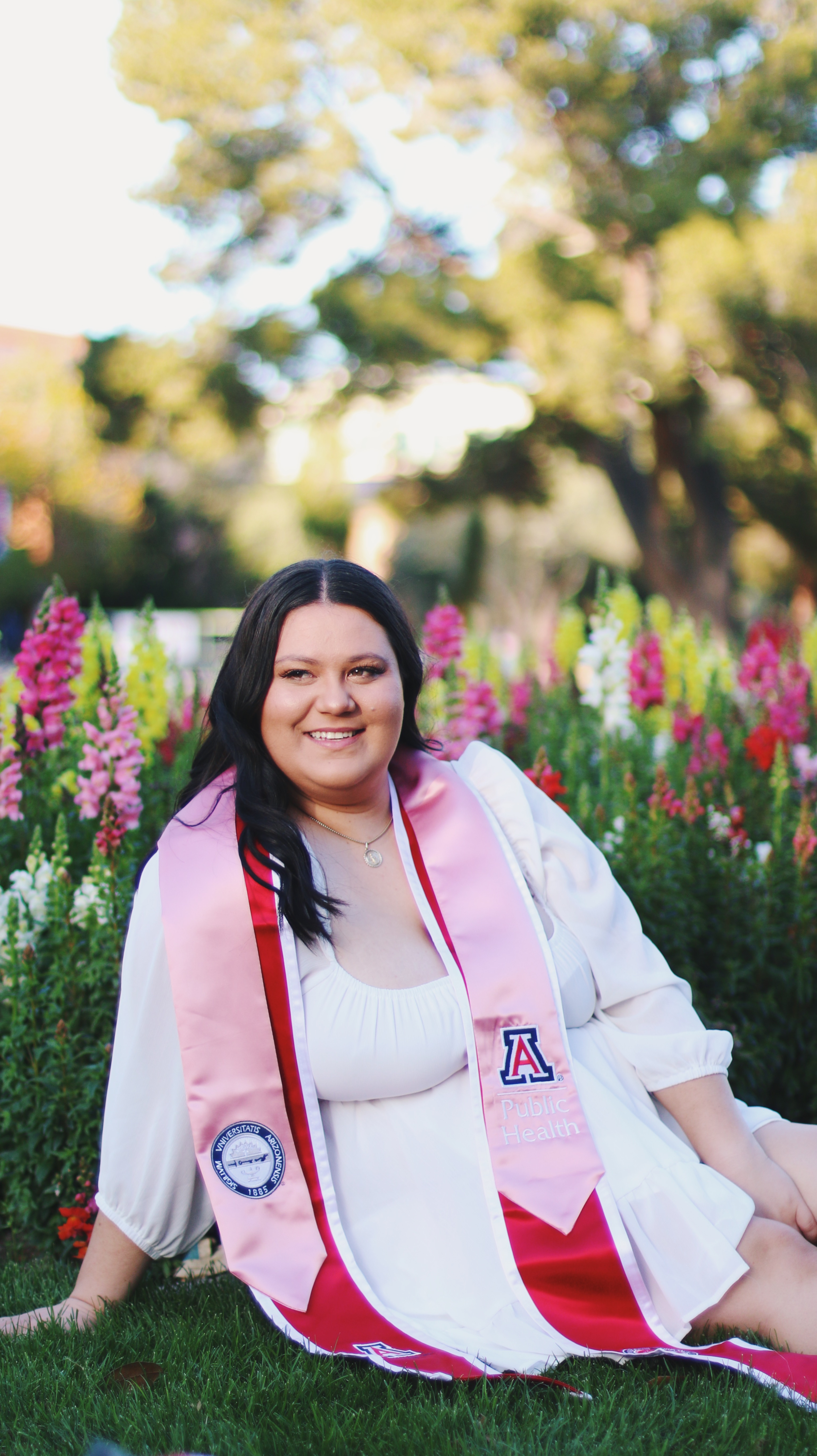 Graduate in white dress with University of Arizona Public Health pink sash sitting in a colorful flower garden