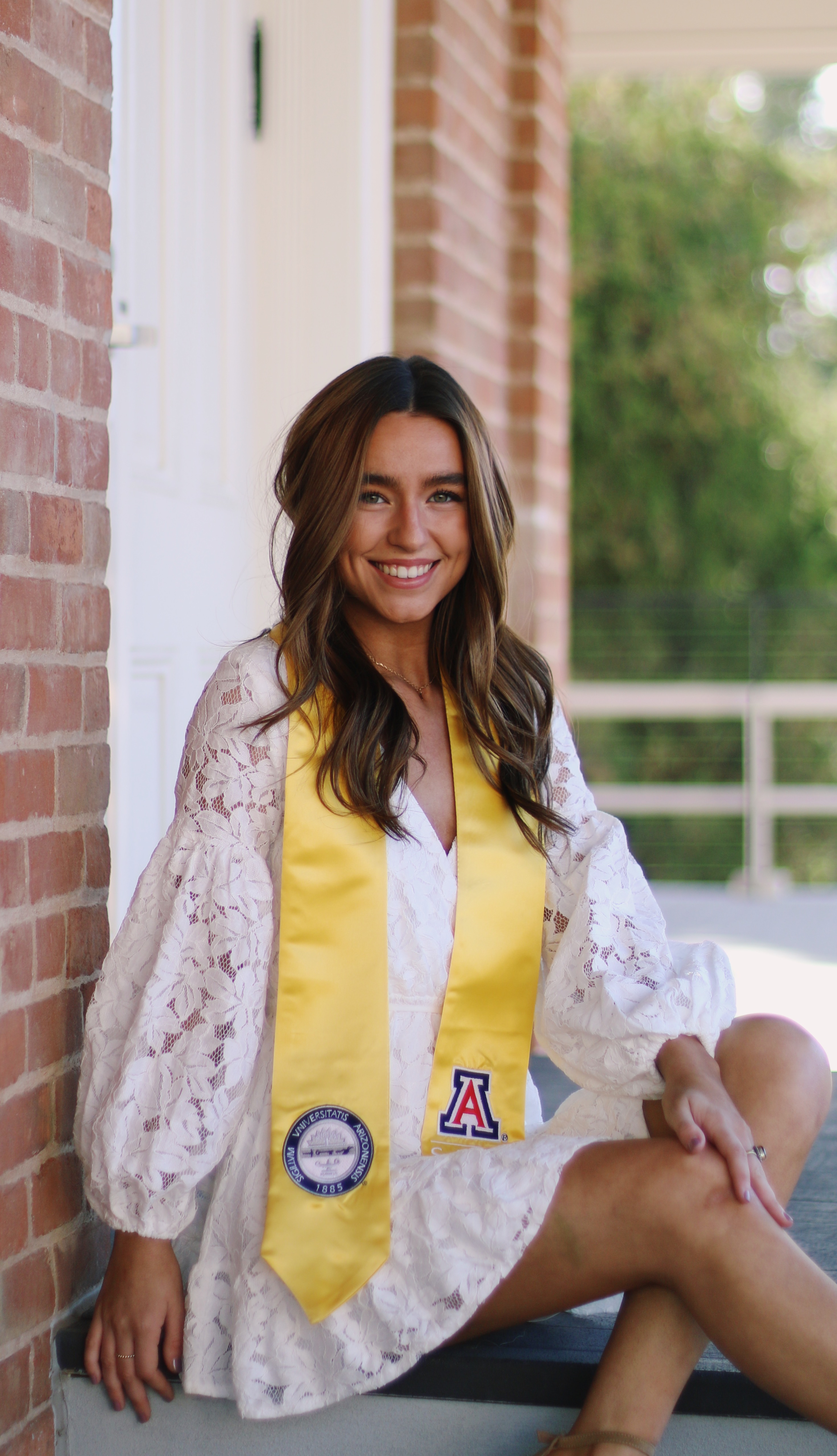 Graduate in white lace dress and yellow University of Arizona sash sitting on steps against a brick wall smiling