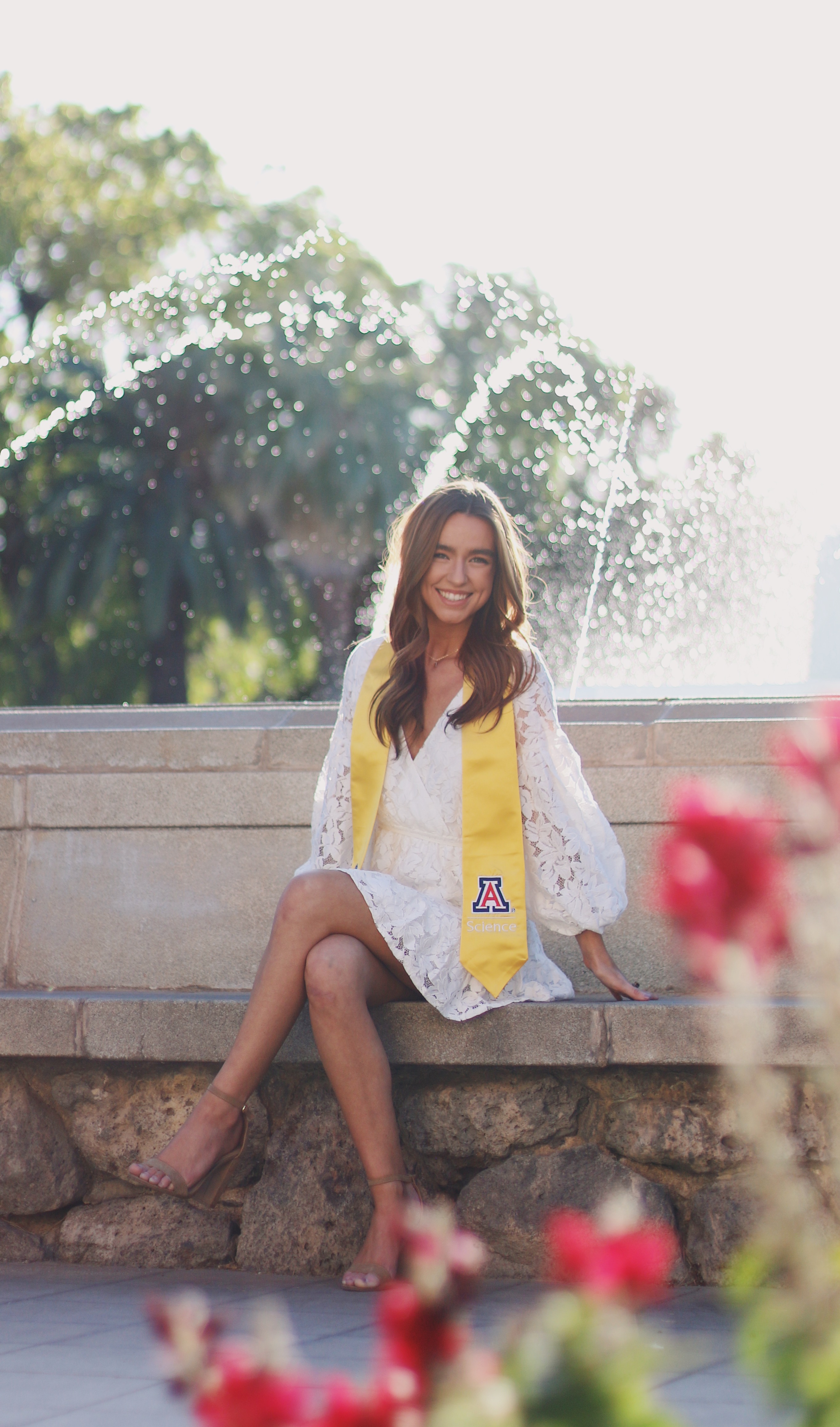 Graduate in white lace dress and yellow University of Arizona sash sitting by a fountain with flowers in the foreground