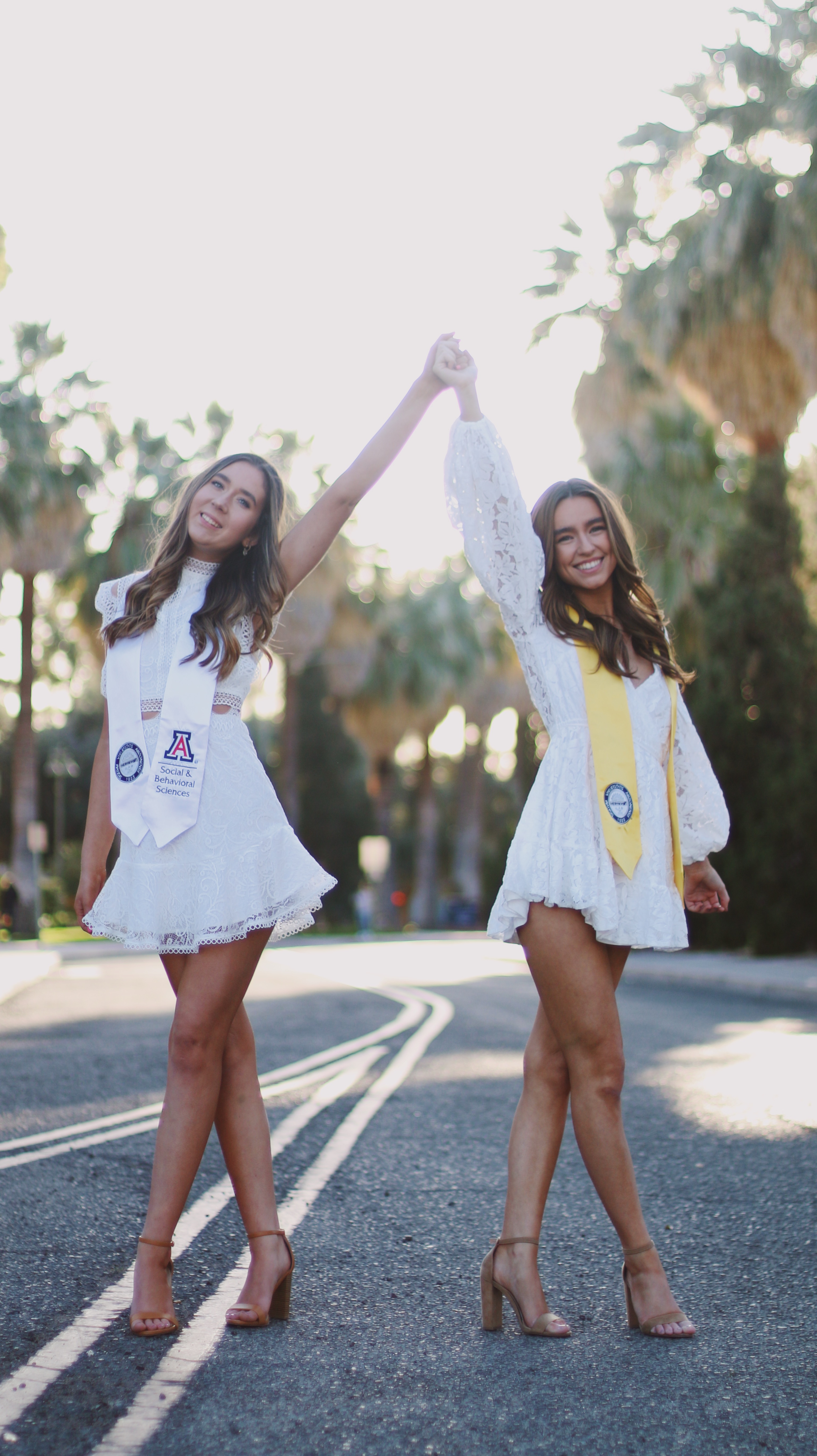 Two female graduates in matching white lace dresses with University of Arizona sashes holding hands raised on a palm tree lined road