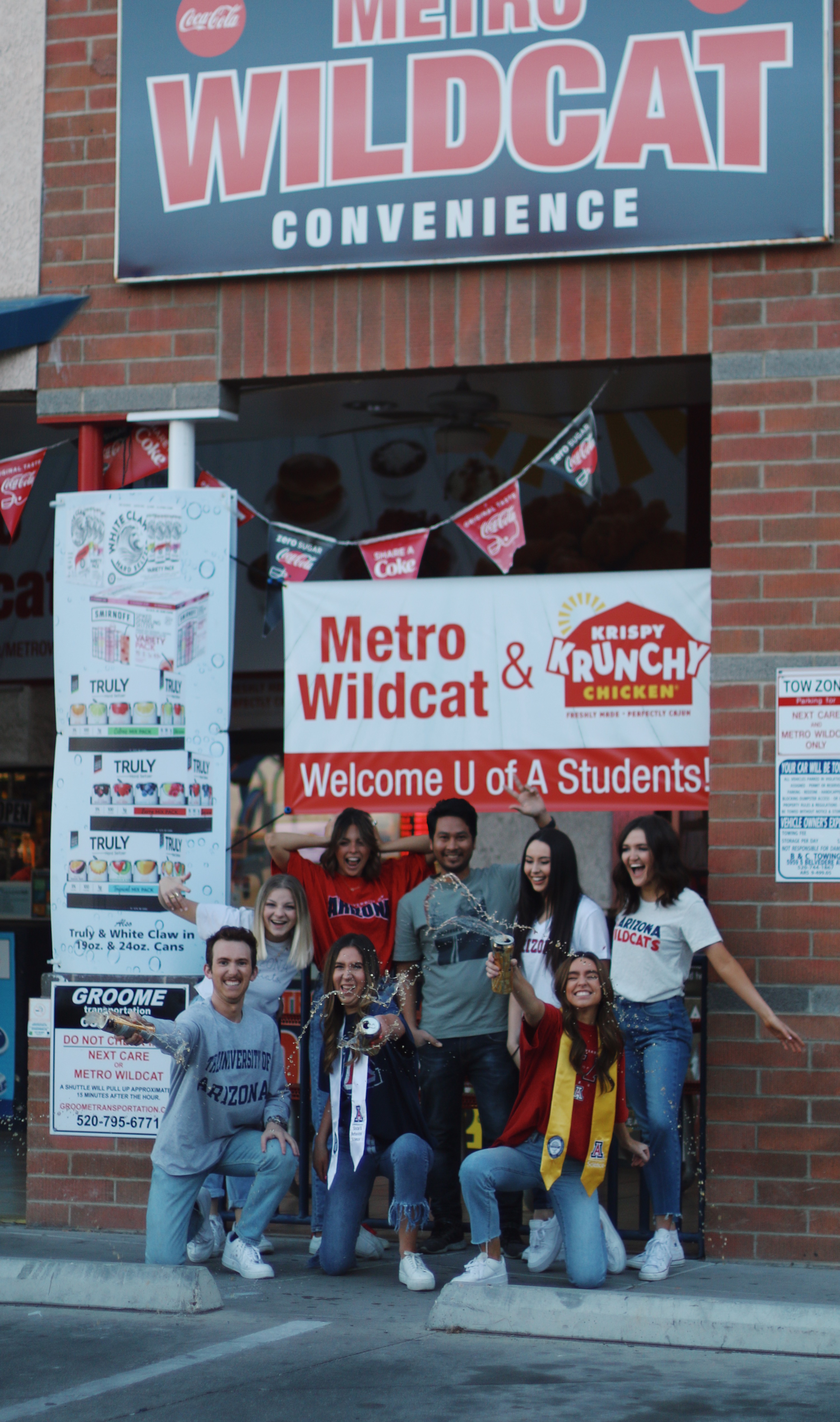 Group of University of Arizona students in school gear celebrating with champagne spray in front of Metro Wildcat convenience store