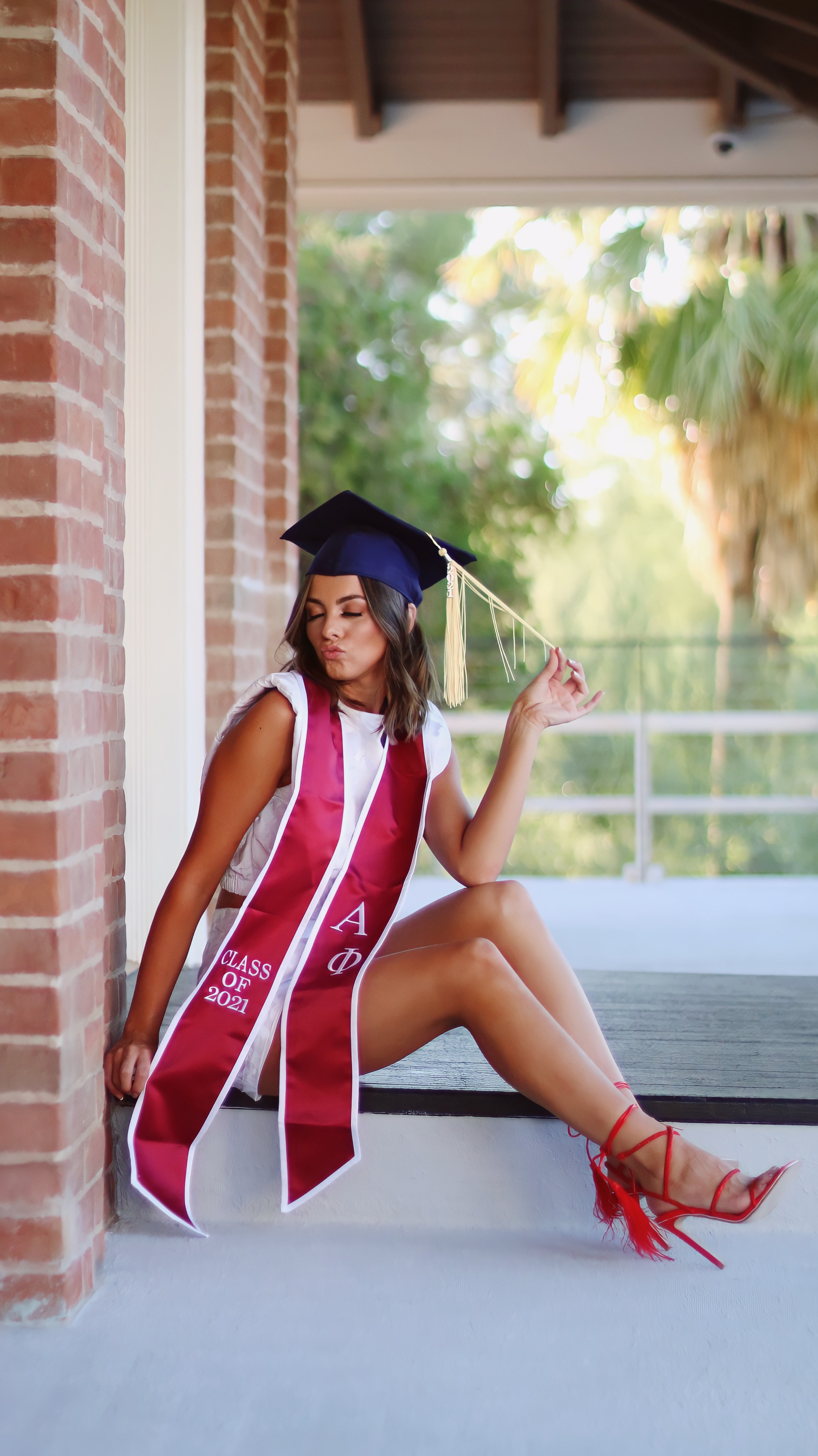 Graduate in navy cap with Alpha Phi Class of 2021 sash sitting on steps blowing a kiss while holding her tassel