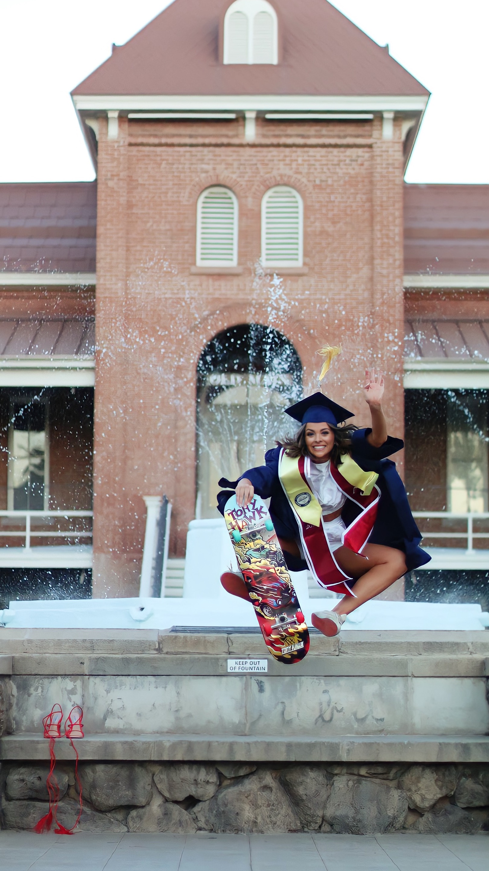 Graduate in navy cap and gown jumping with a Tony Hawk skateboard in front of the Old Main fountain