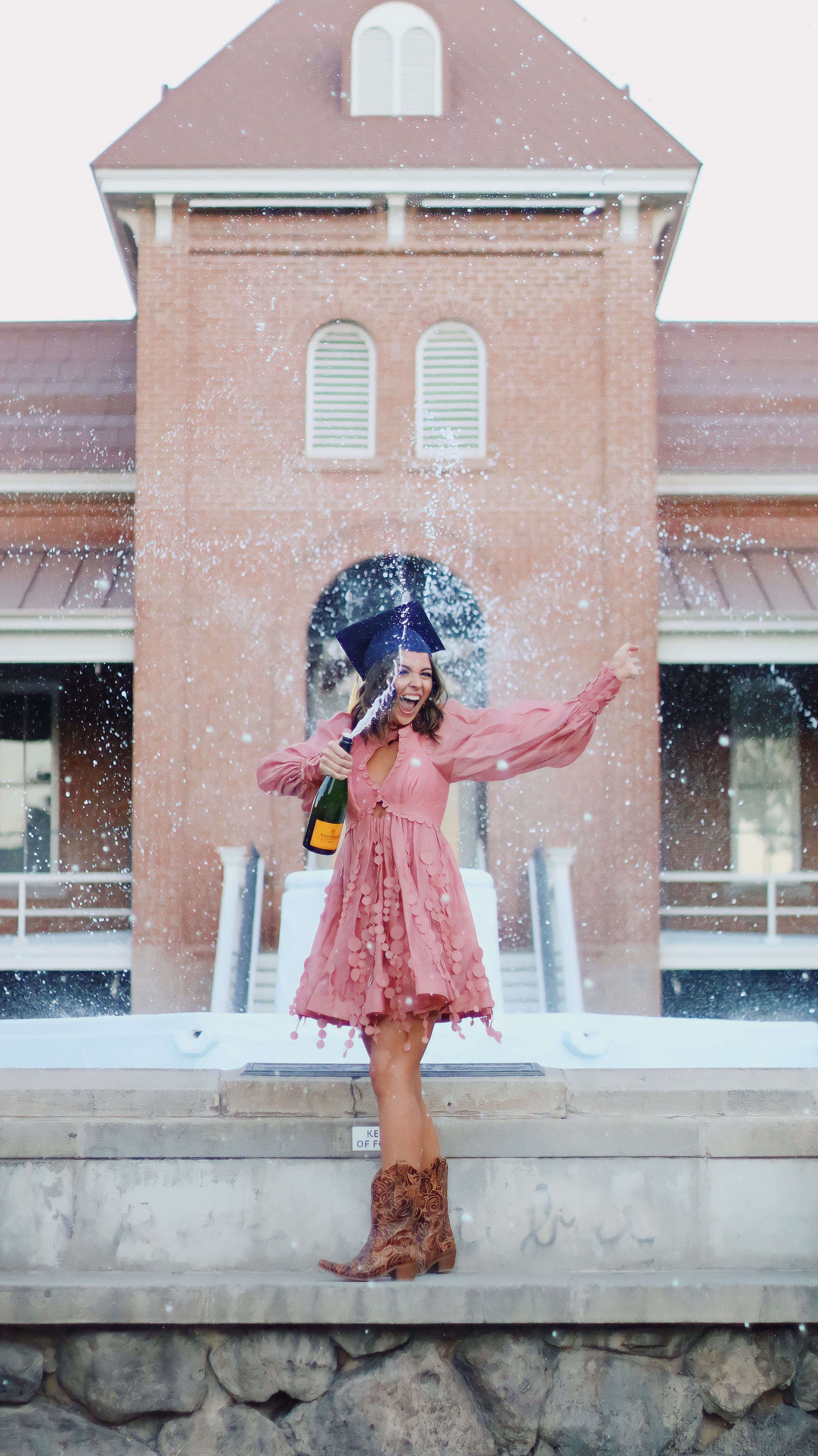 Graduate in pink dress and cowboy boots popping champagne in front of a historic brick building
