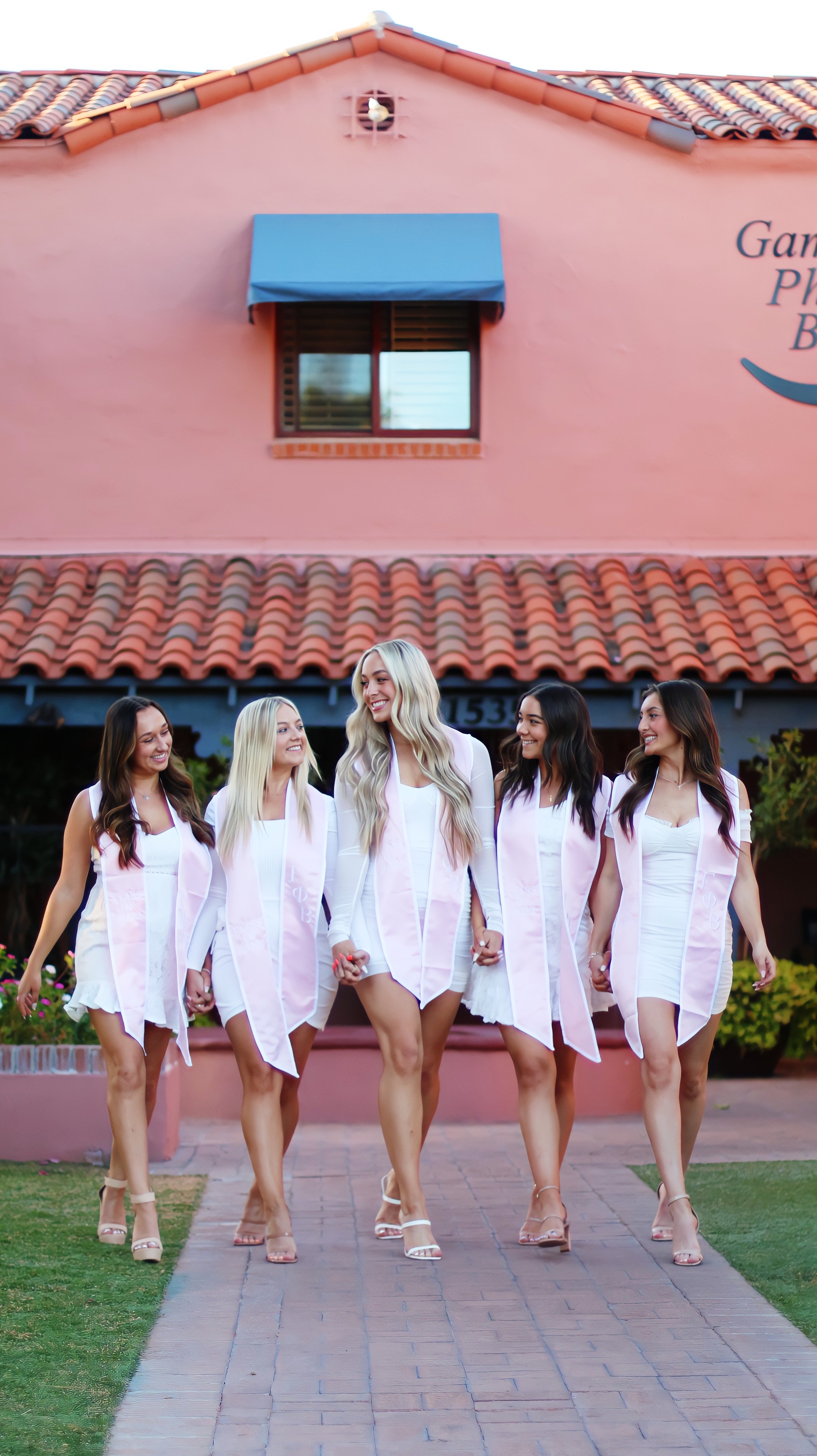 Group of graduates in white dresses walking in front of a brick building with their arms around each other