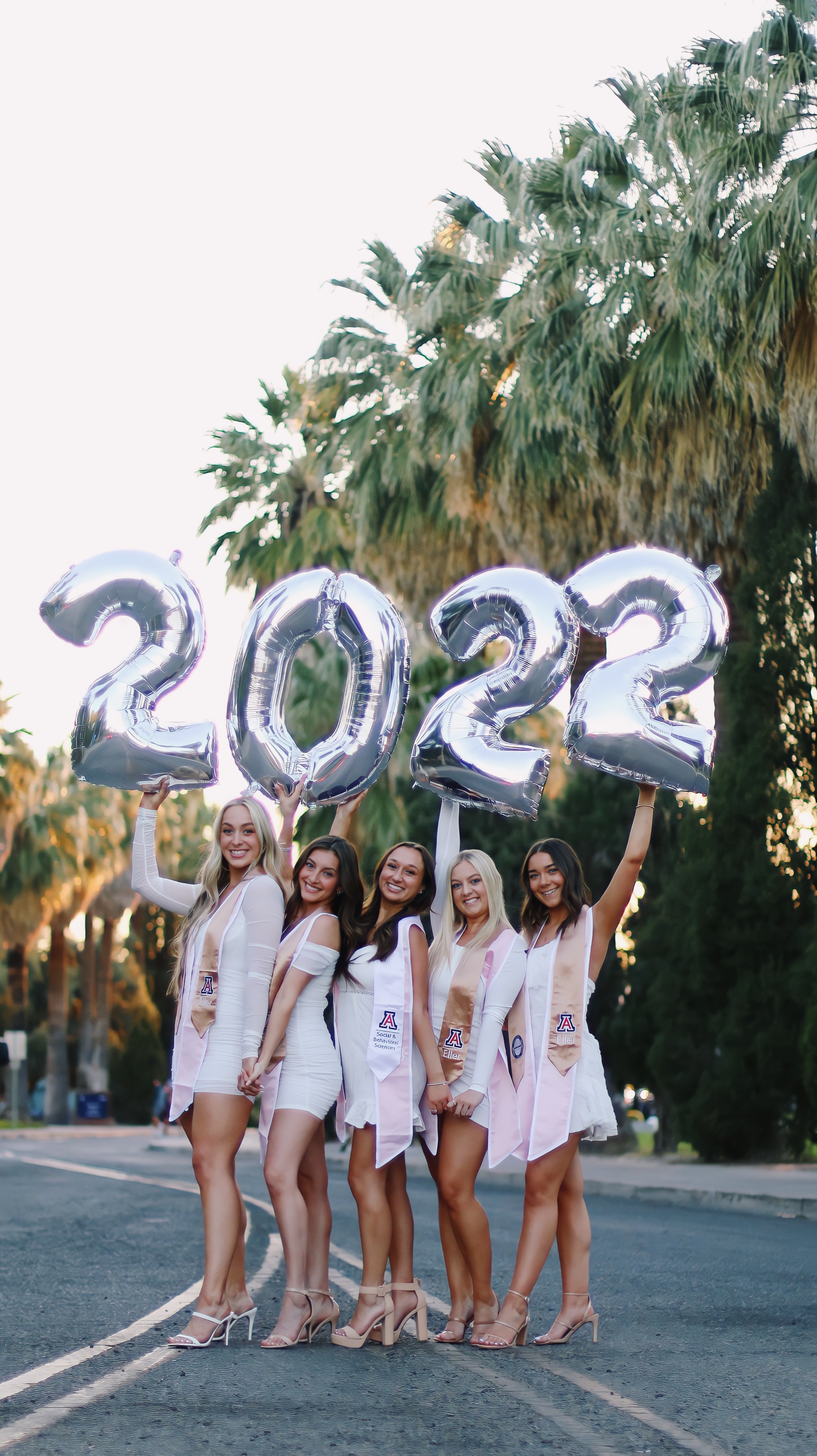 Group of graduates in white dresses holding up silver 2022 balloons
