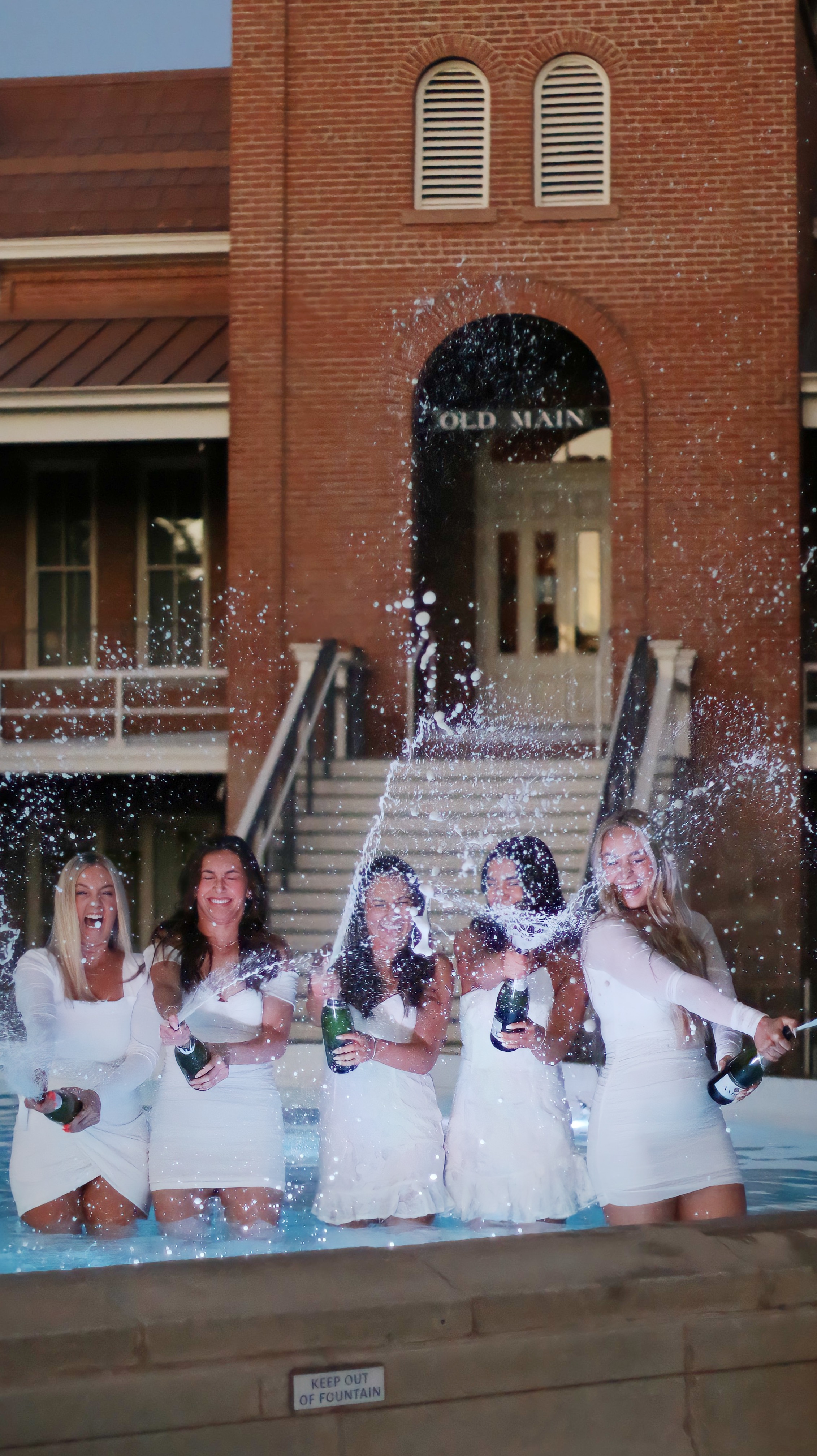 Group of graduates in the white dresses standing in the fountain popping champagne bottles in the air
