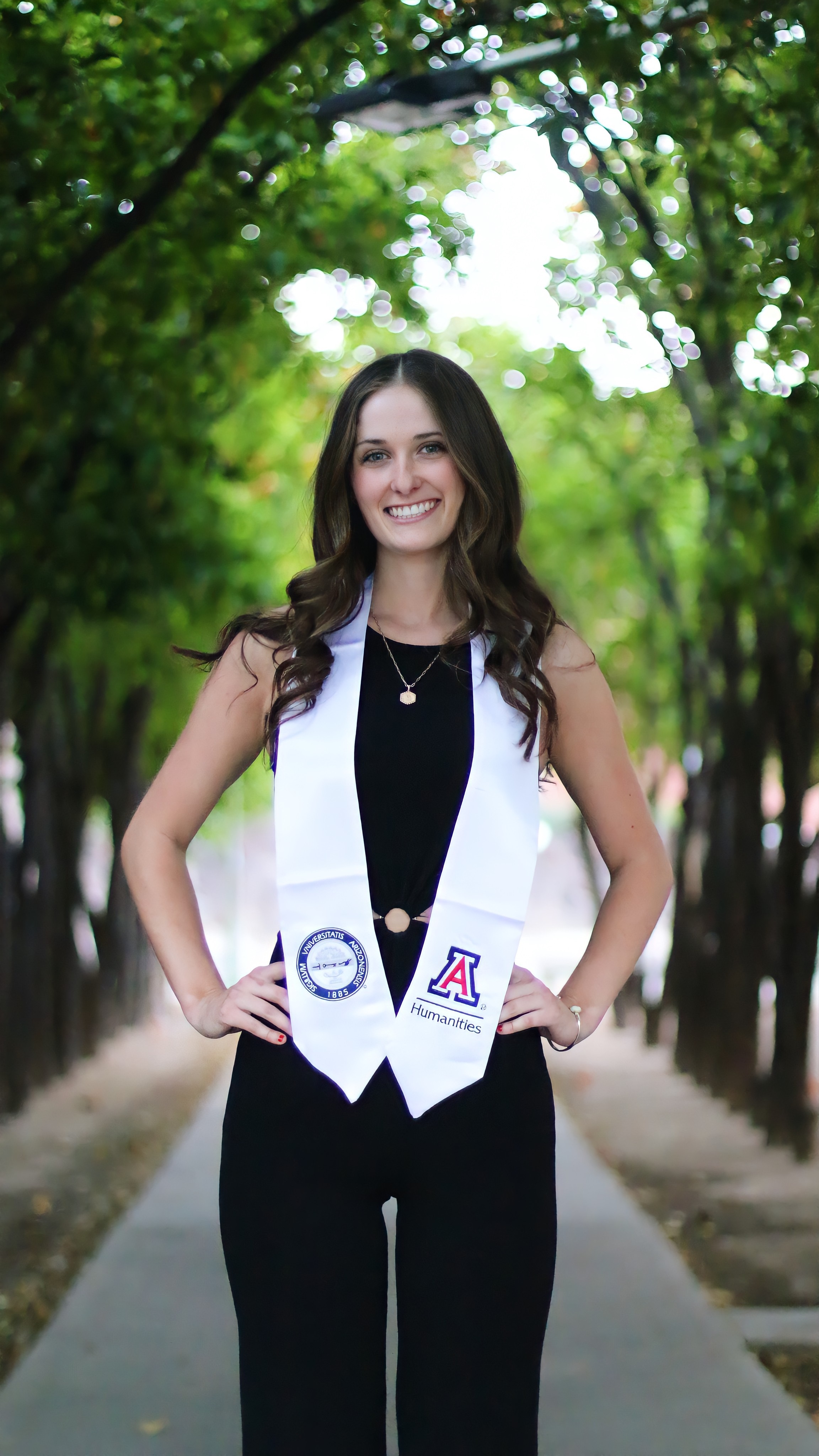 Young graduate in a black jumpsuit with university sash on standing in front of a row of trees