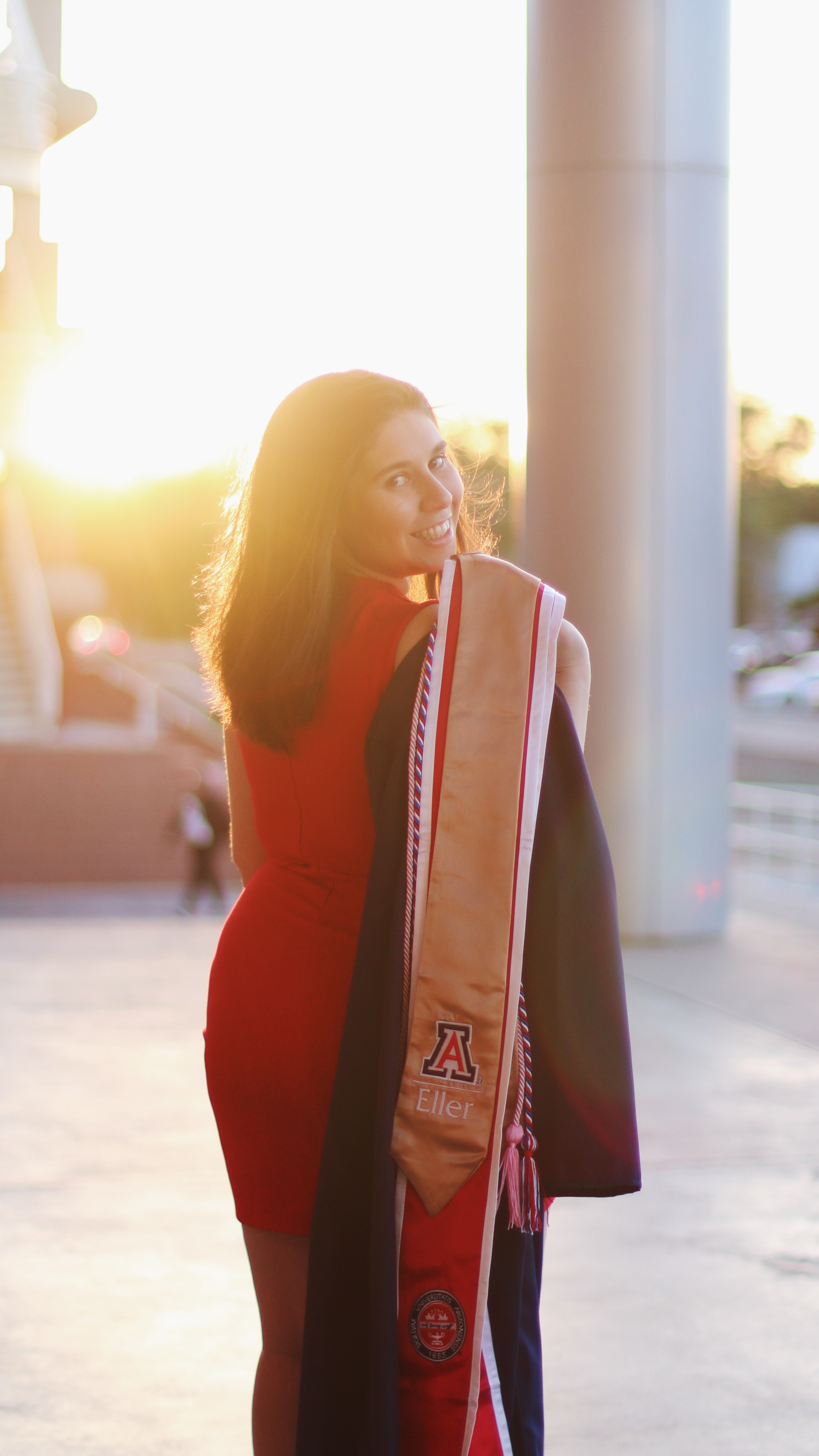 Youg women in red dress and university gown and sash looking back at the camera with the sunset in the background