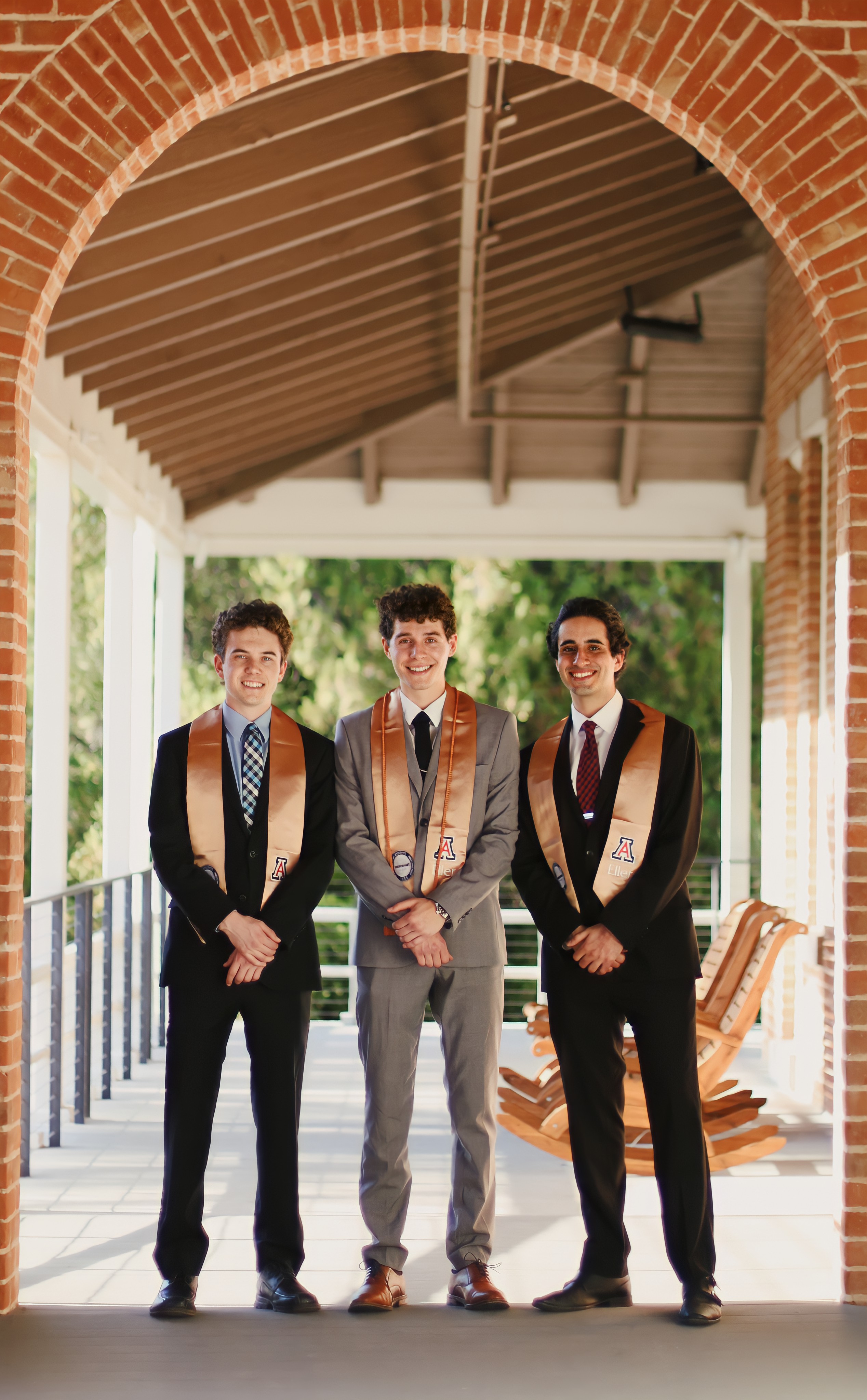 Group of graduate men standing in a row under a brick building porch with their arms crossed and looking at the camera