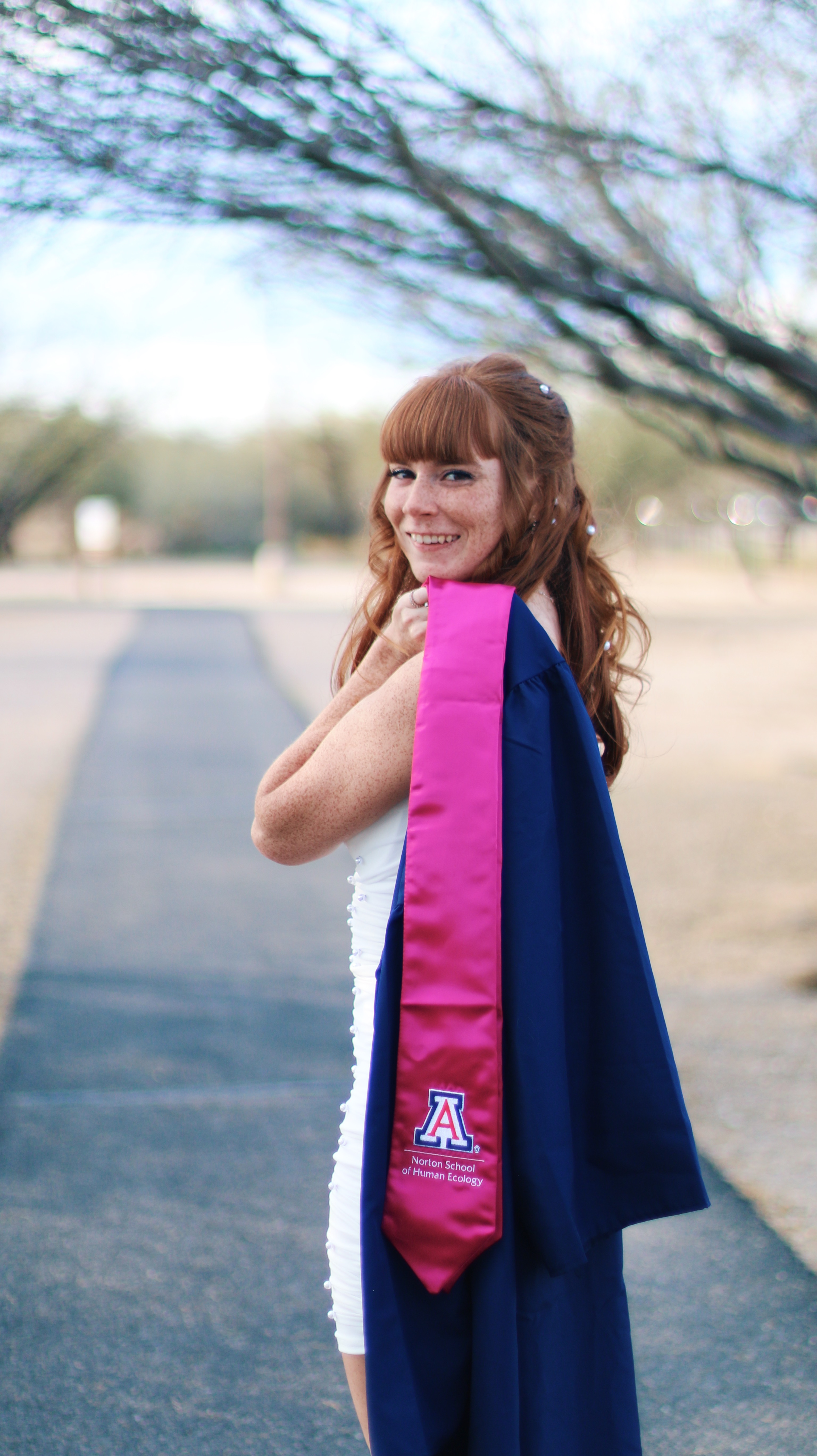 Graduate walking on a road looking back at the camera with the university sash and gown over her shoulder