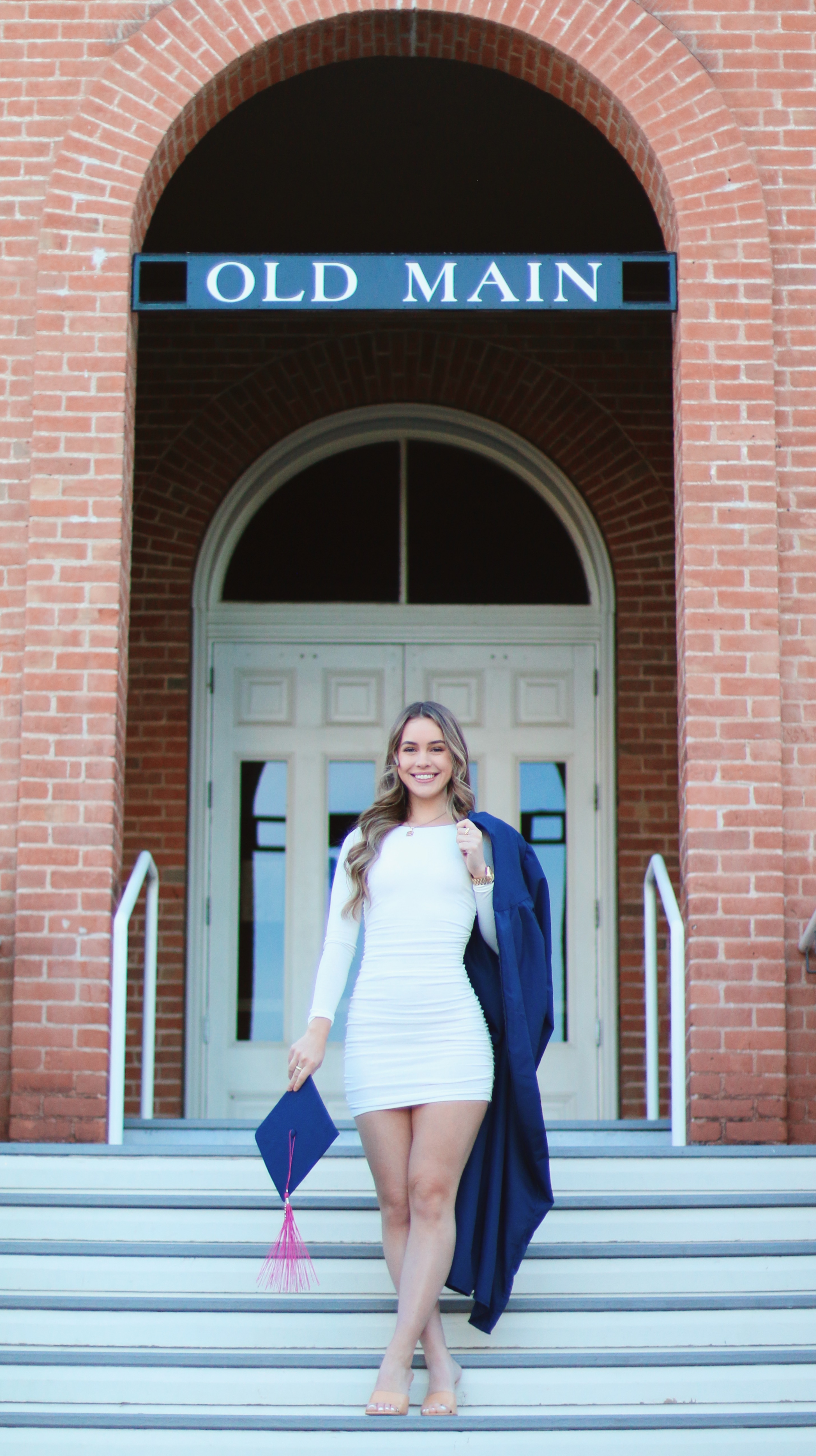 Graduate in white dress and University of Arizona sash sitting on the steps of a brick building smiling at the camera