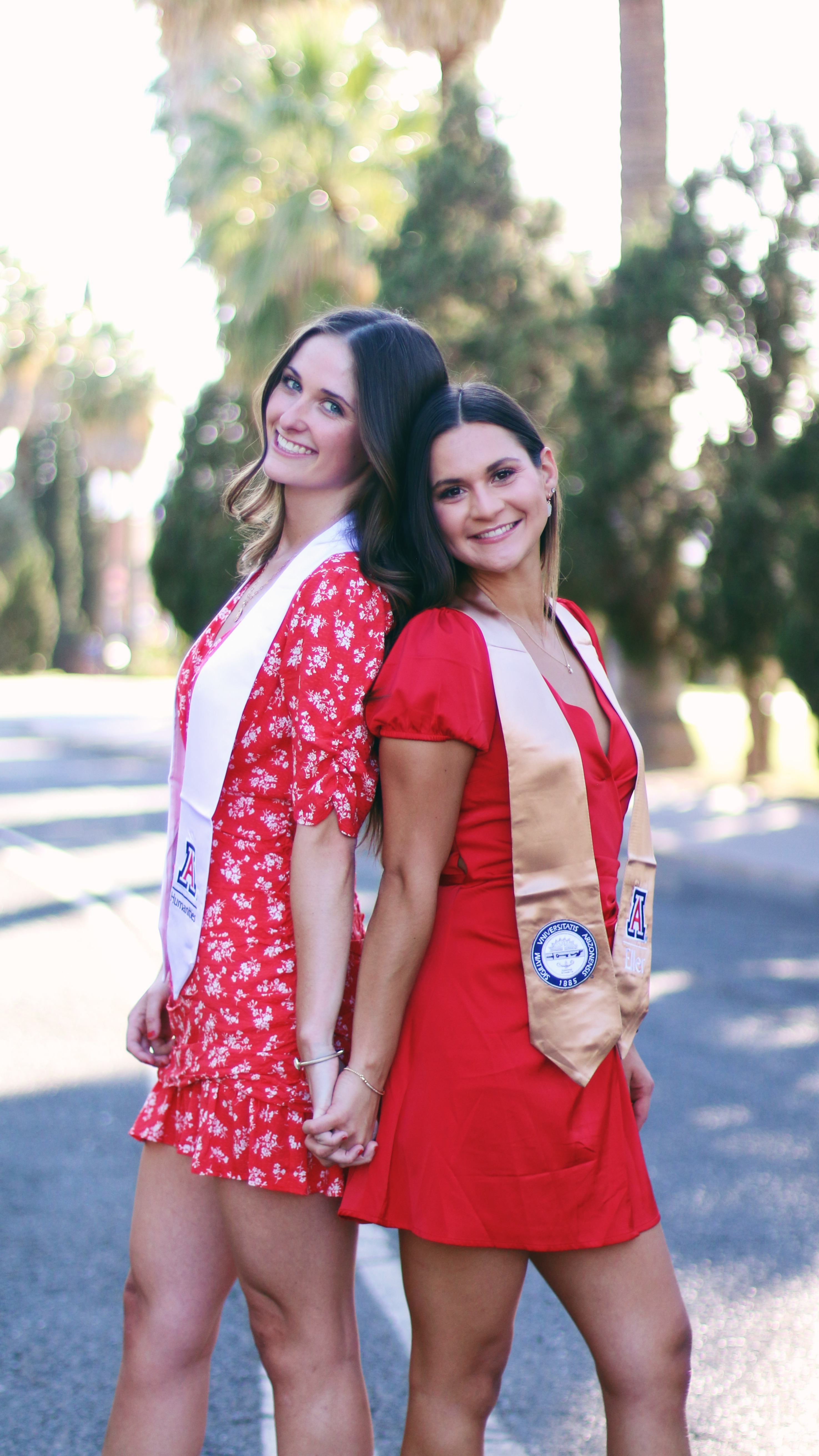 Two young women wearing red dresses and their university sashes smiling at the camera while posing back to back